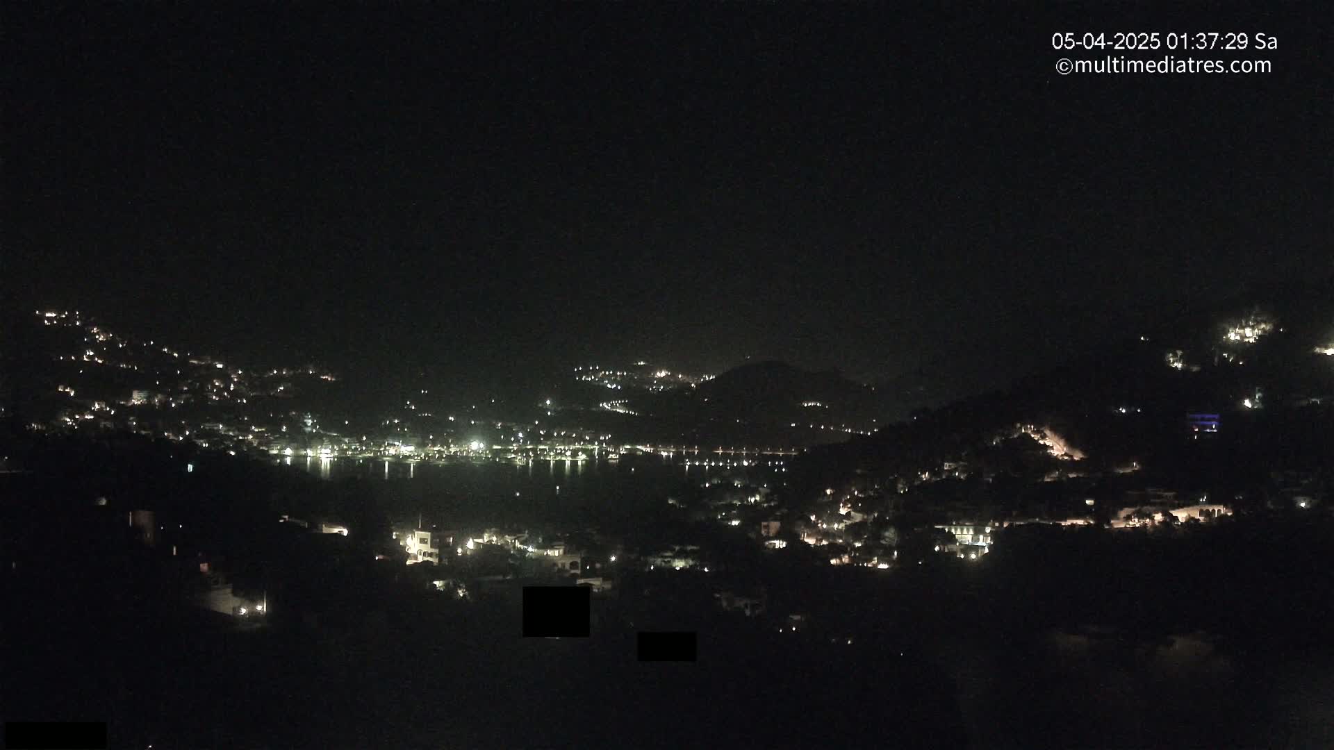 A nighttime view of a coastal town, with hills dotted with lights, overlooking a dark body of water under a clear sky.