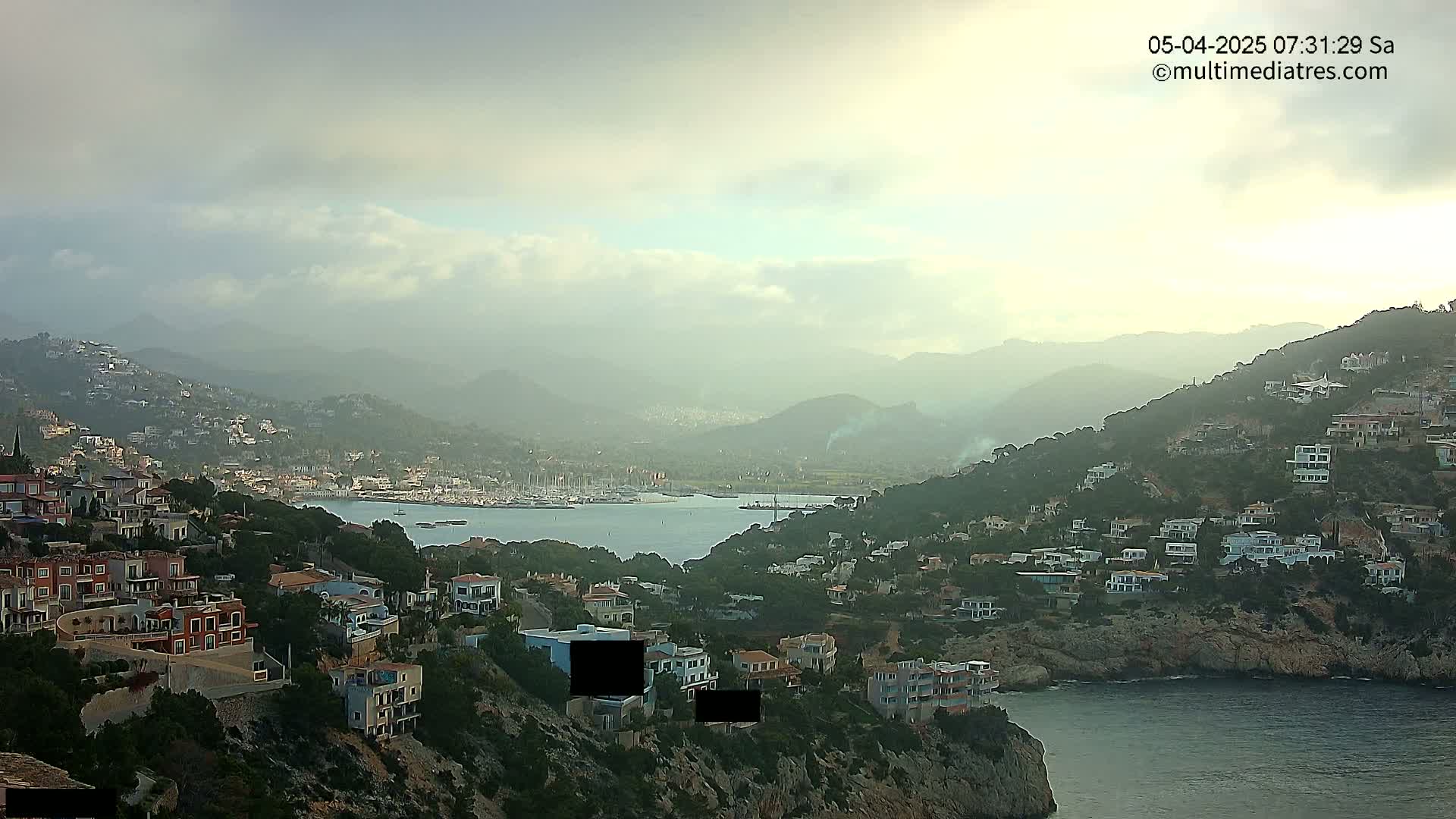 A hazy, overcast morning reveals a coastal town nestled on hillsides overlooking a calm bay filled with boats.