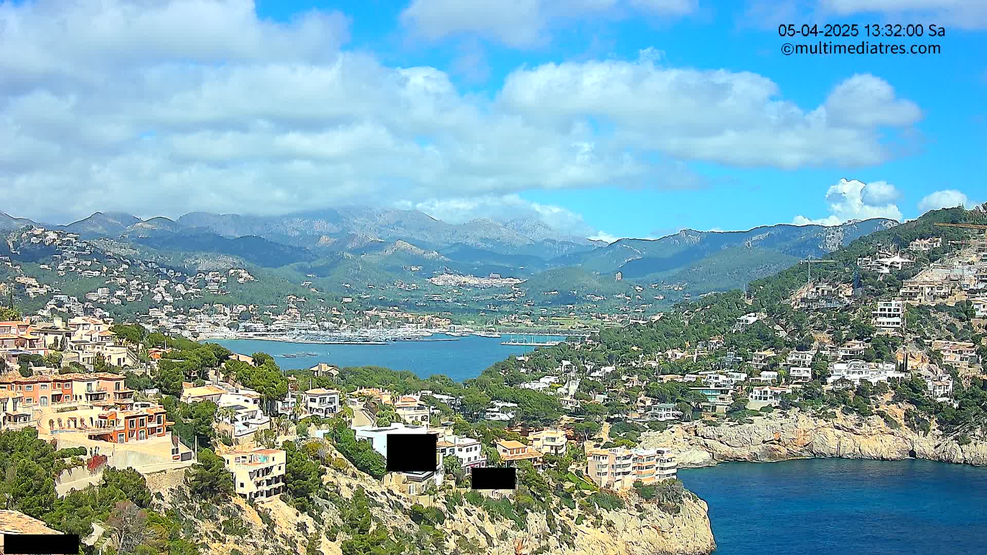 A coastal town nestled on a hillside overlooking a calm bay, with mountains in the background under a partly cloudy sky.