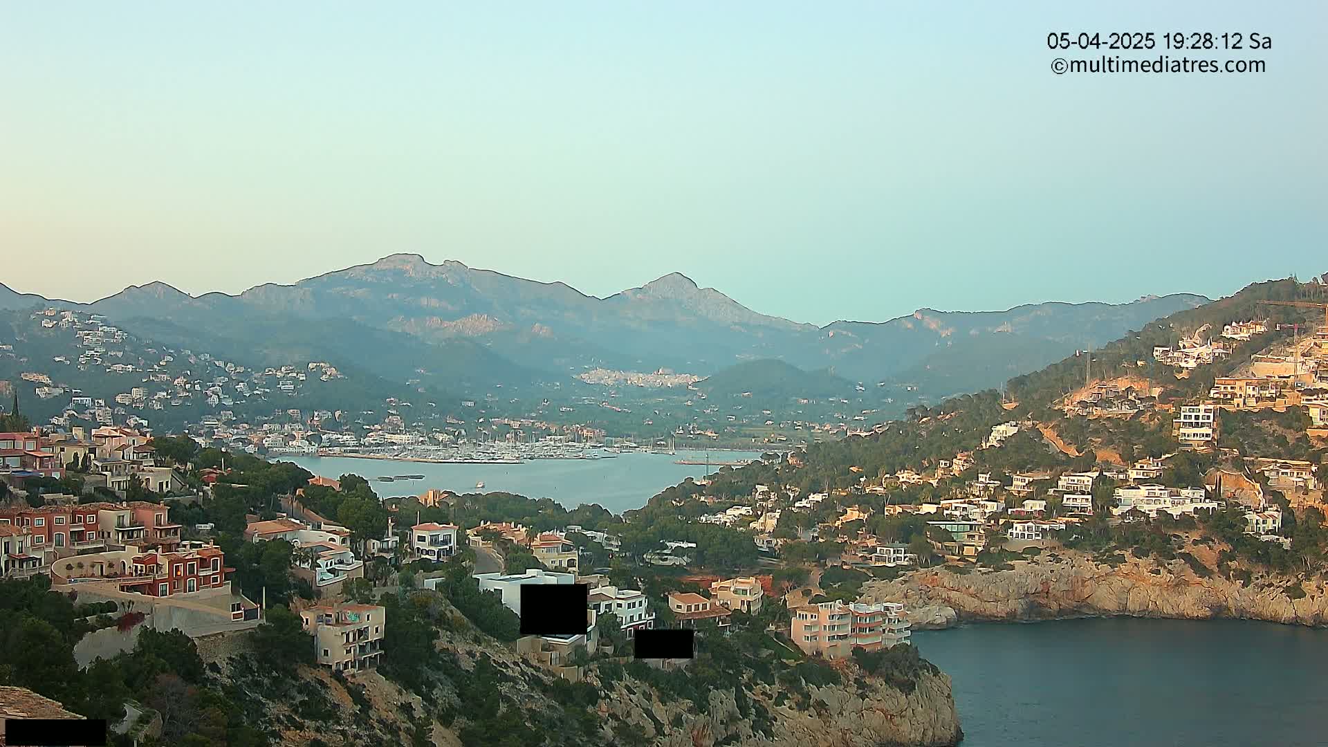 A coastal town nestled on a hillside overlooking a calm bay with a marina, backed by a range of mountains under a clear sky.