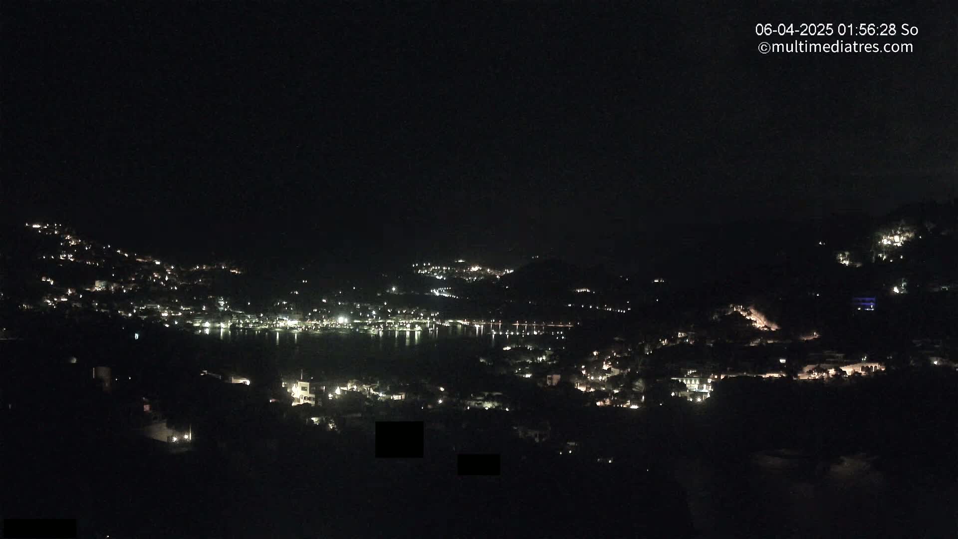 A nighttime view shows a coastal town with scattered lights on hillsides overlooking a dark body of water under a clear, dark sky.
