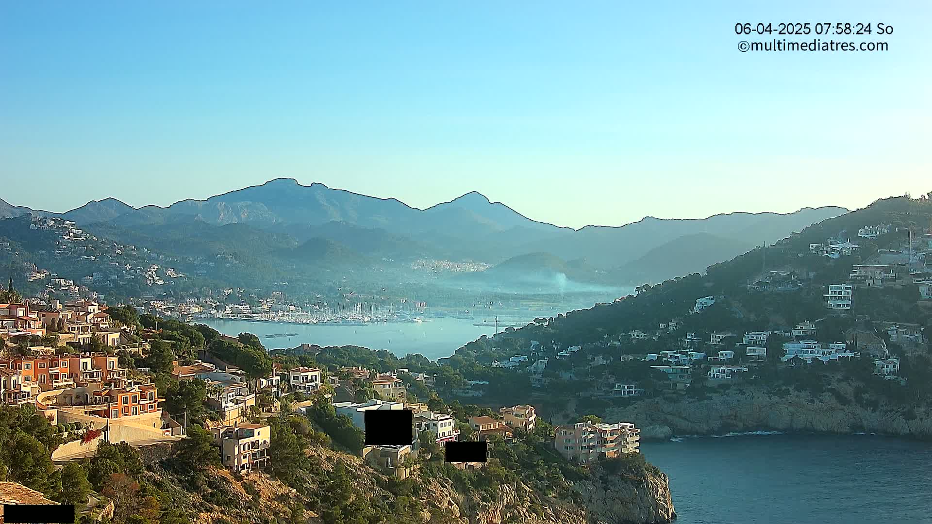 A clear, sunny day overlooking a coastal town nestled between a calm bay and a mountain range, with numerous houses built into the hillside.