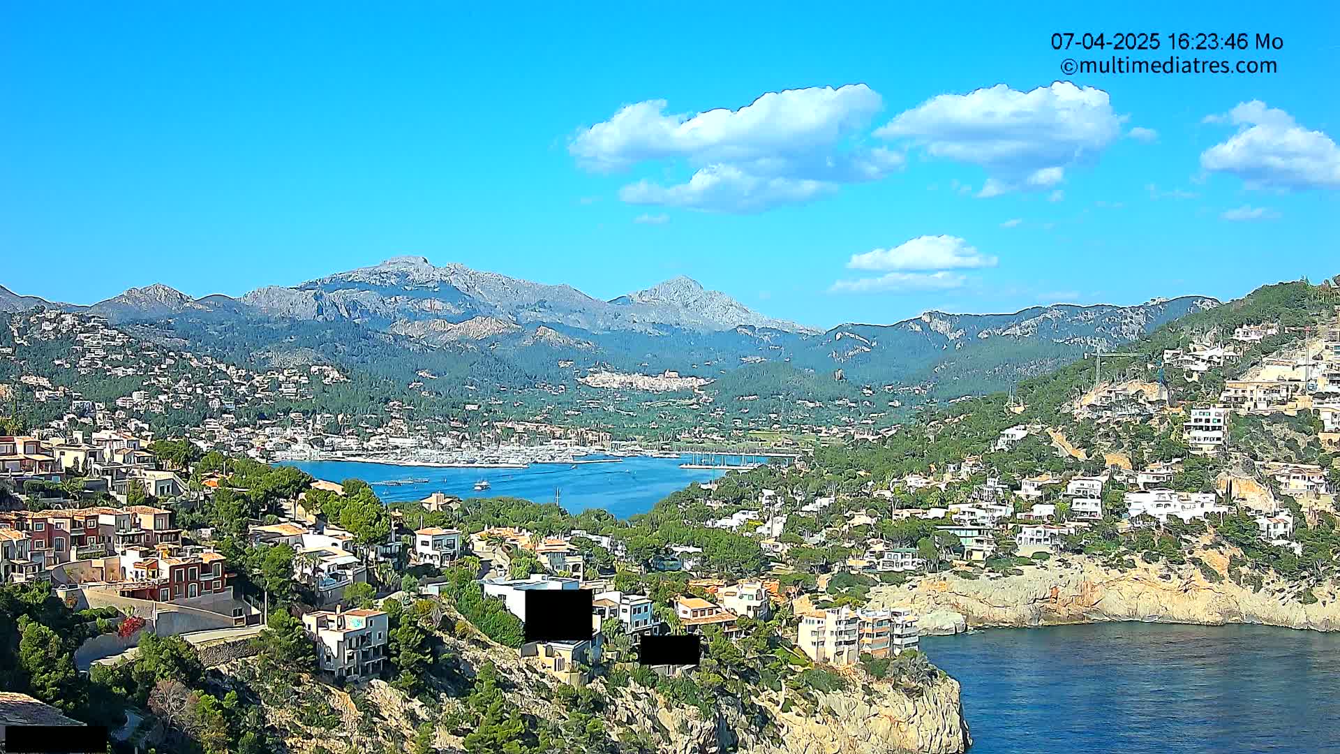 A coastal town nestled in a mountainous region, under a mostly sunny sky with a few scattered clouds, overlooks a calm bay filled with boats.