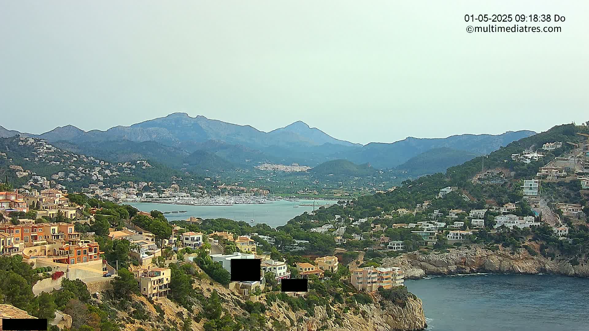 A hazy coastal town nestled on a hillside overlooking a calm bay filled with sailboats, with mountains in the background.