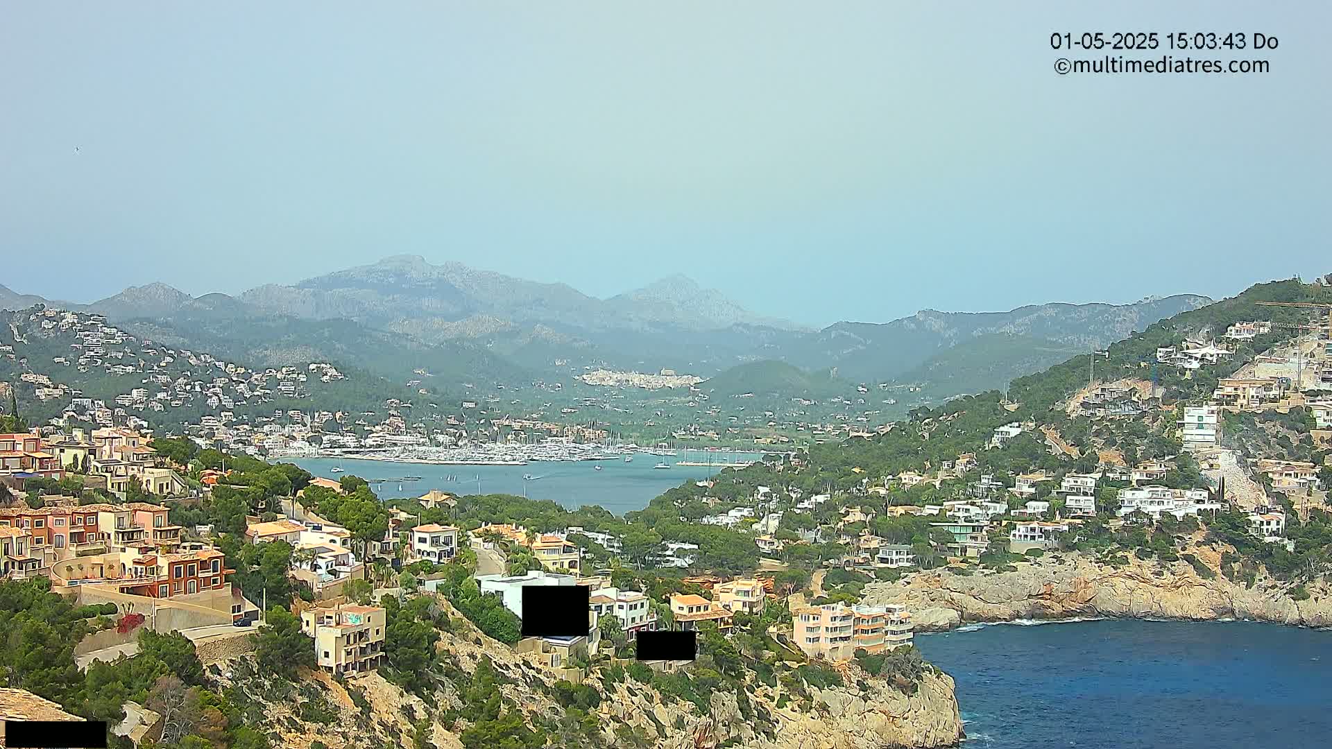 A hazy, sunny day overlooks a coastal town nestled in hills, with a harbor filled with boats and mountains in the background.