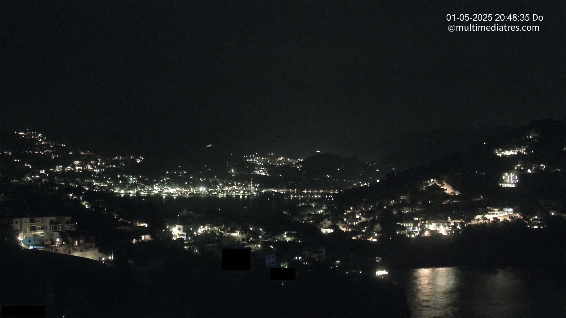 A nighttime view of a coastal town nestled on hillsides, with scattered lights illuminating buildings and the dark water below.