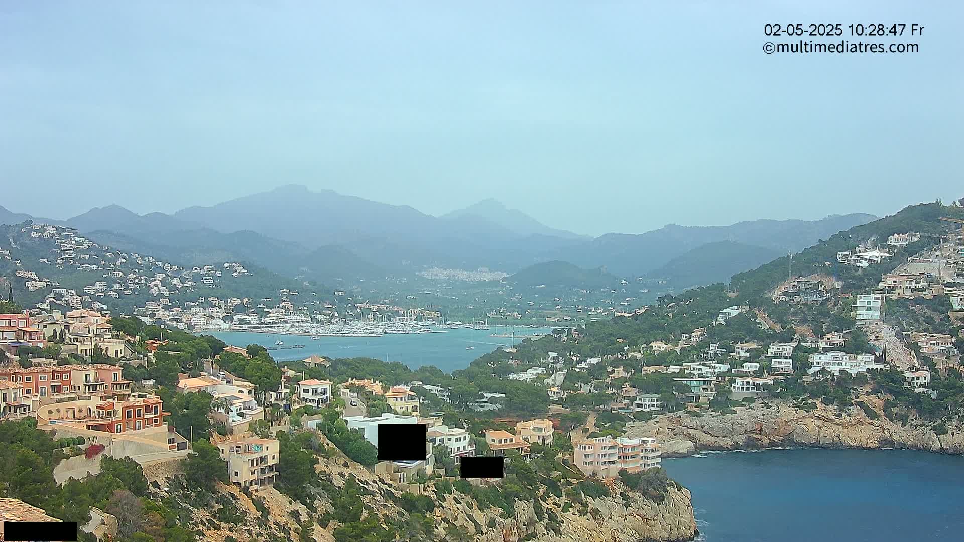 A hazy-looking coastal town with many white houses is nestled amongst green hills and overlooks a calm bay filled with boats, all under a clear sky.