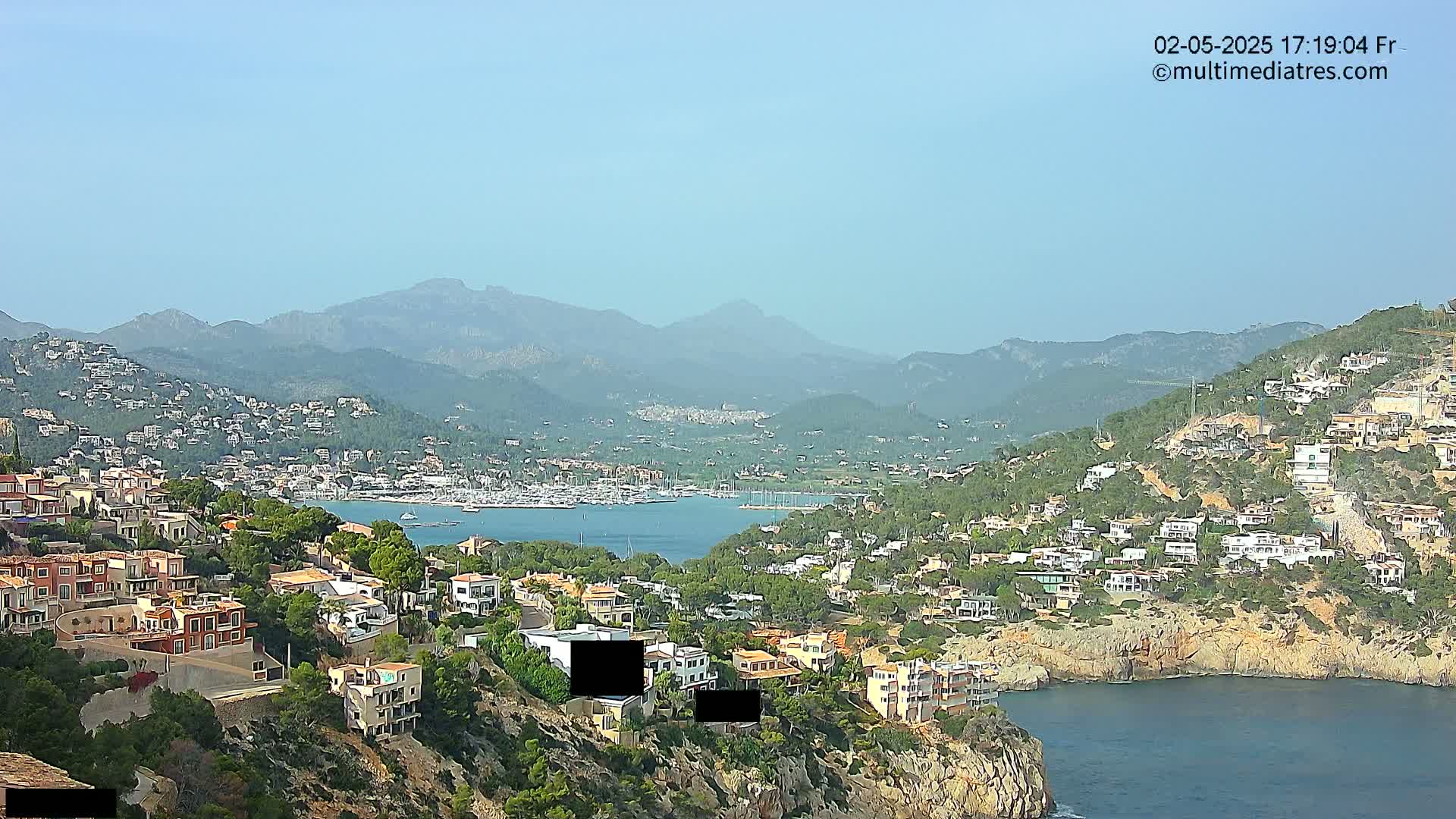 A coastal town nestled on a hillside overlooks a calm bay filled with boats, with mountains in the background under a clear, sunny sky.