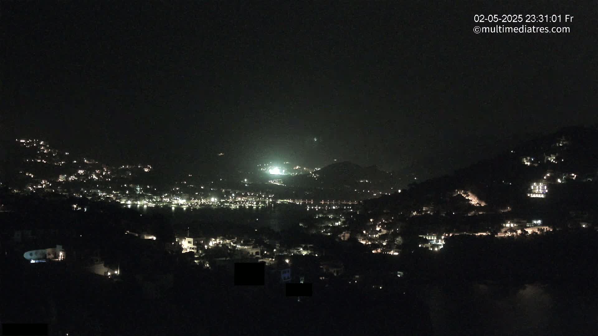 A nighttime view of a coastal town nestled on hillsides, illuminated by scattered lights, under a clear, dark sky.