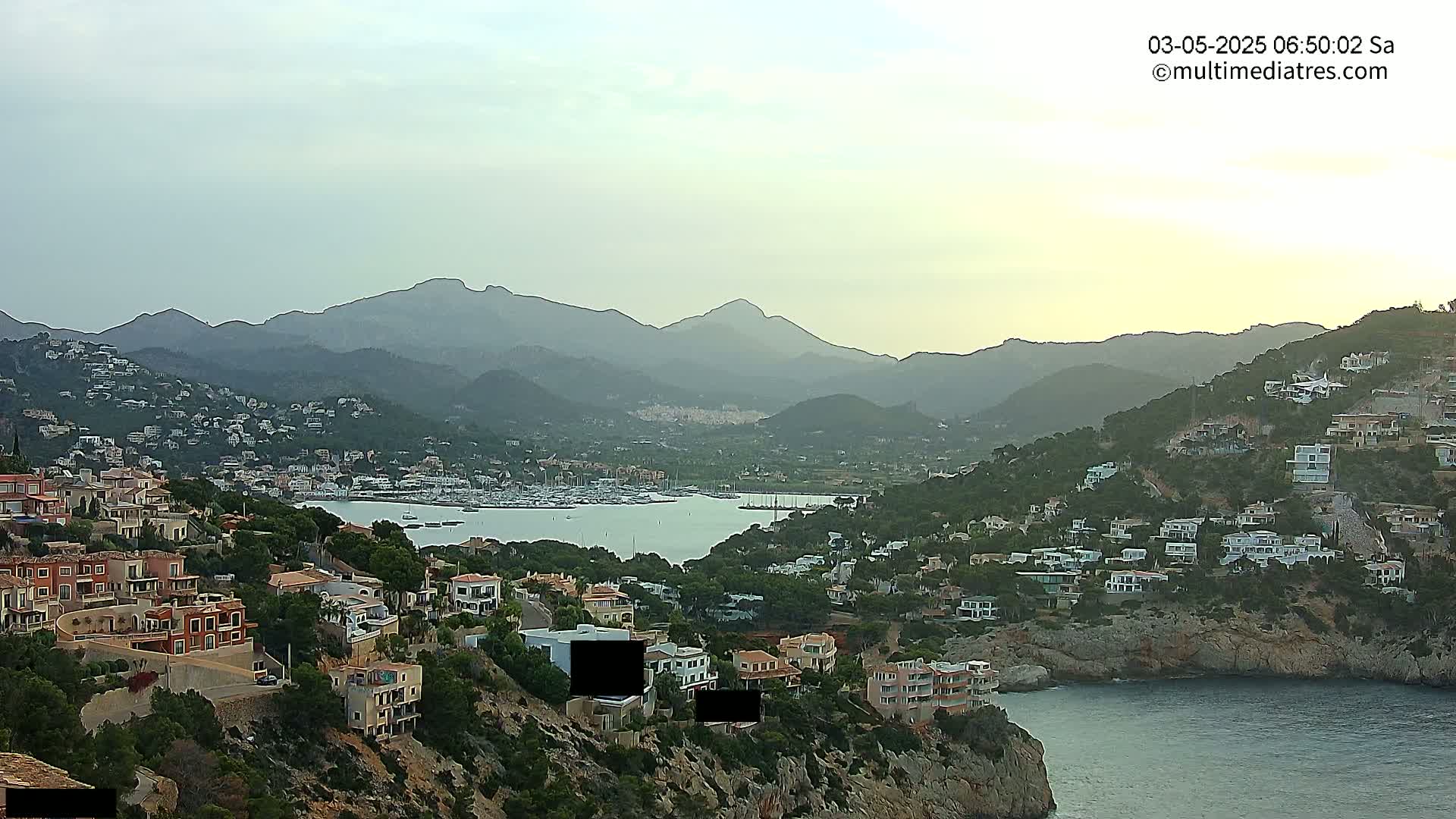 A coastal town nestled into hills overlooking a calm bay with numerous boats, all under a hazy, overcast sky.