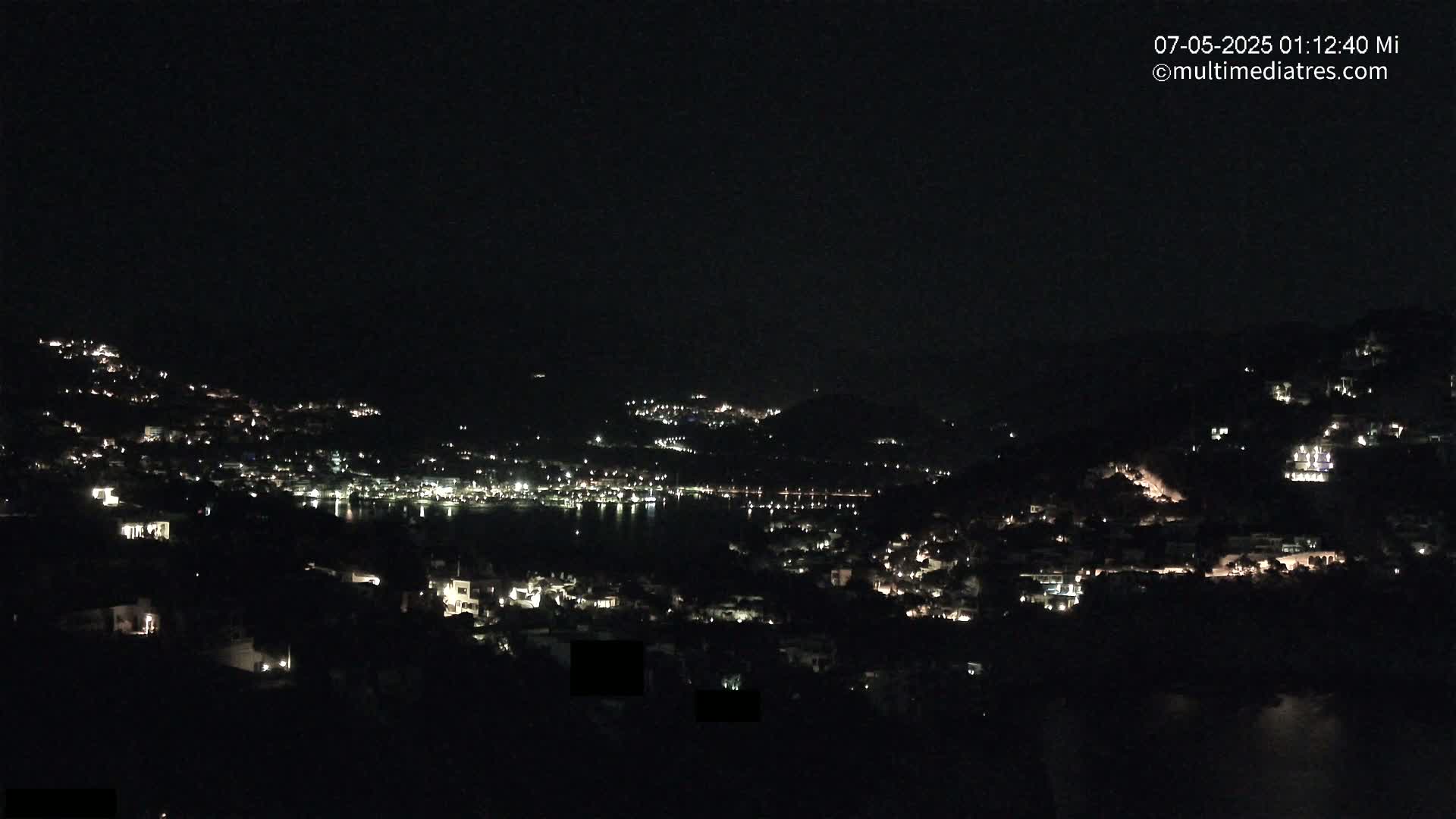 A nighttime view of a coastal town illuminated by scattered lights, with a dark sky and calm water visible in the distance.