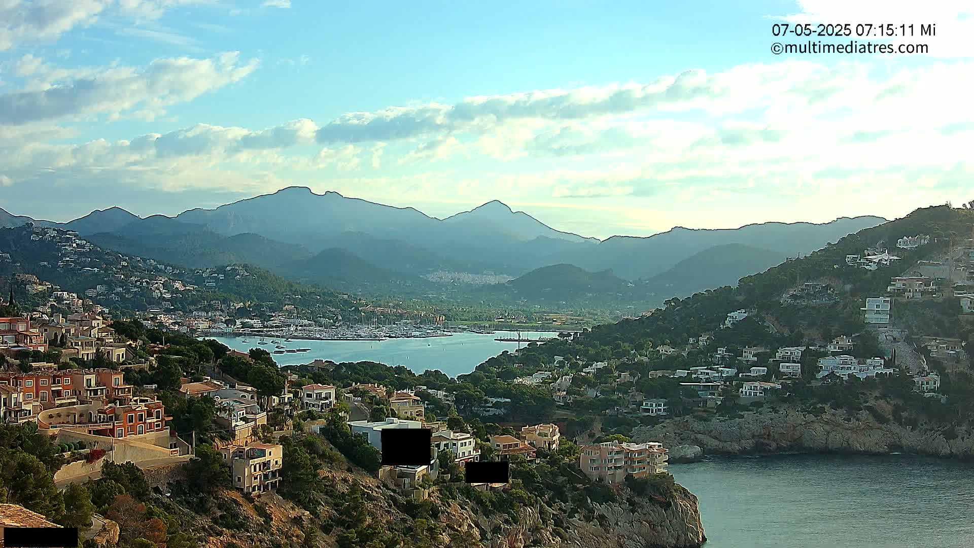 A coastal town nestled against mountains under a partly cloudy sky overlooks a calm bay filled with boats.