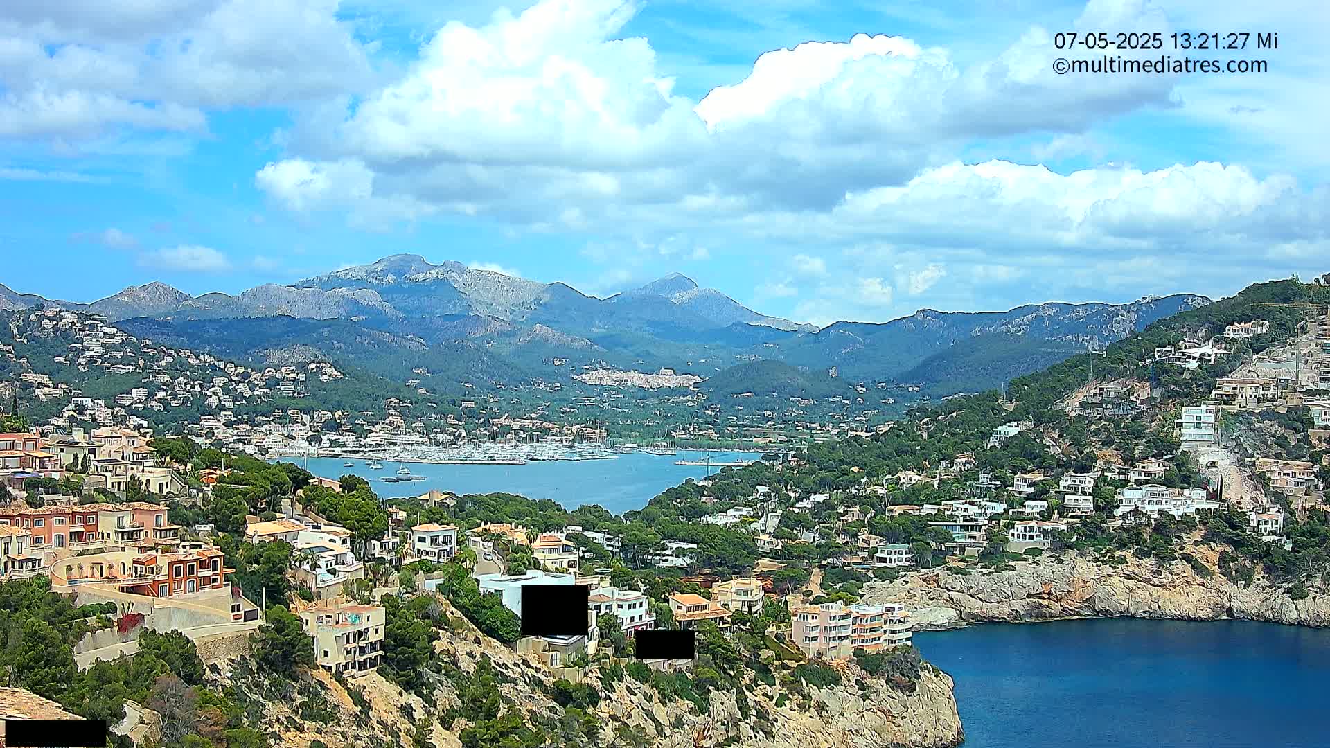A partly cloudy day reveals a picturesque coastal town nestled into a hillside, overlooking a calm bay filled with boats and backed by a range of mountains.