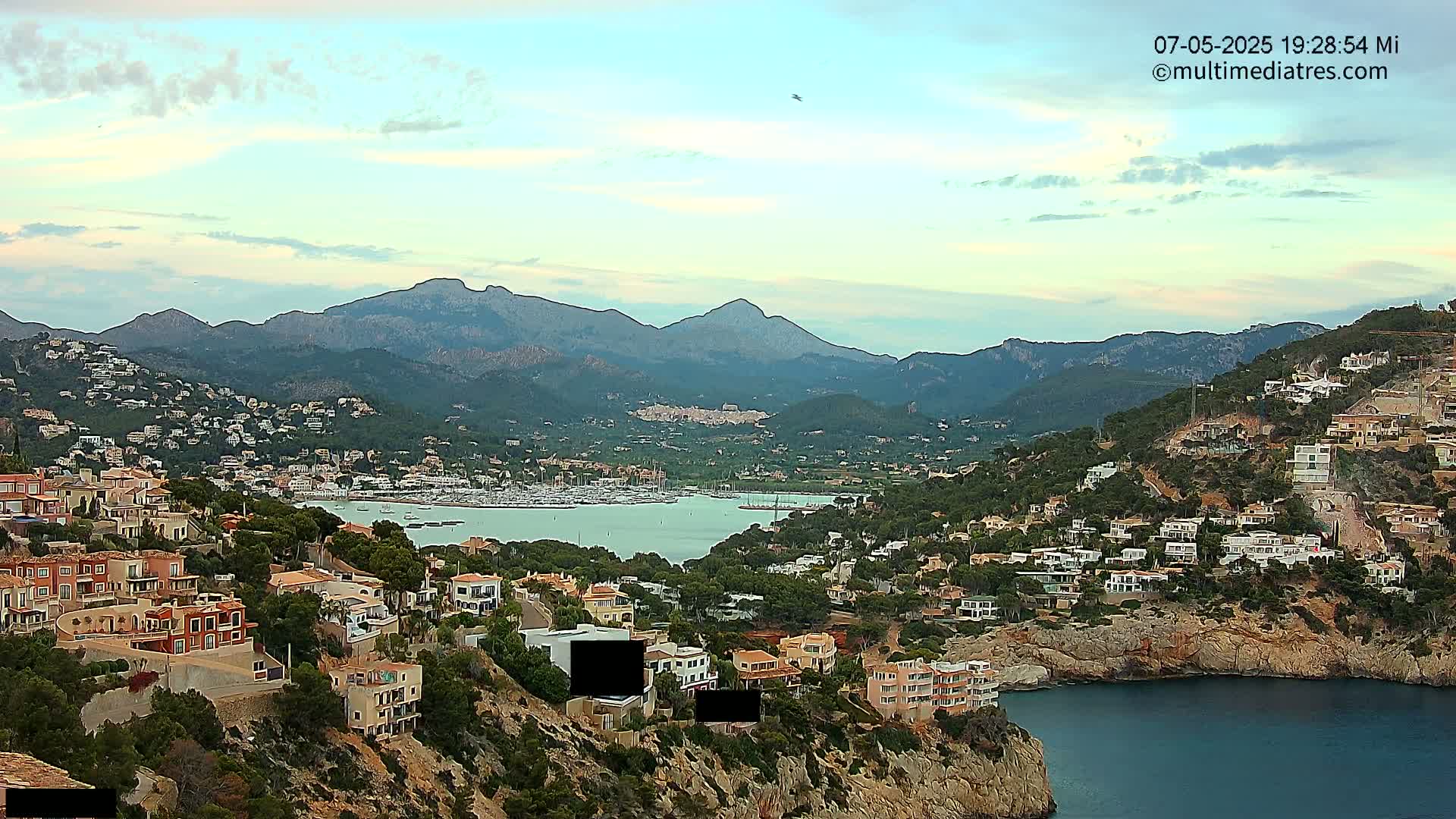 A coastal town nestled in a mountainous region, under a partly cloudy sky, overlooks a calm bay filled with boats.
