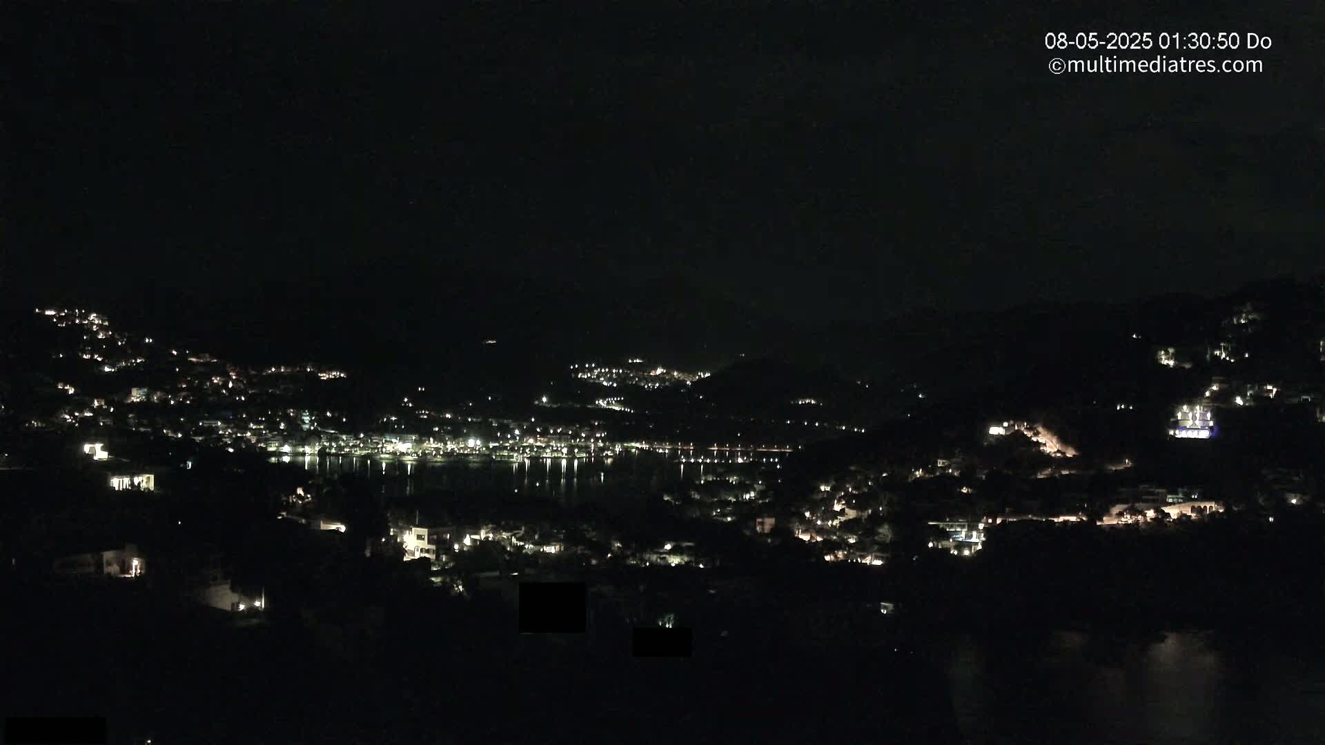 A nighttime view of a coastal town nestled on hillsides, with scattered lights illuminating the buildings and a dark body of water reflecting the lights.
