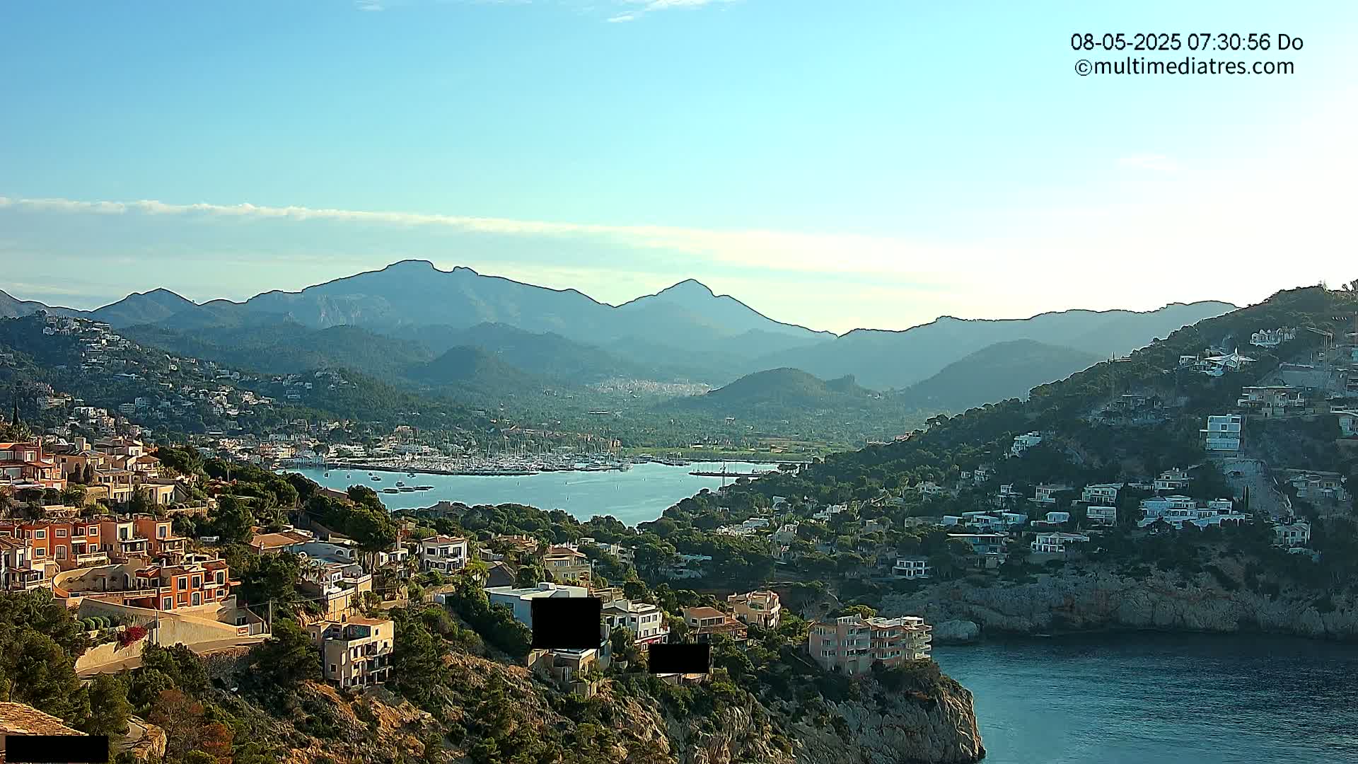 A coastal town nestled between hills and mountains overlooking a calm bay filled with boats under a clear, sunny sky.