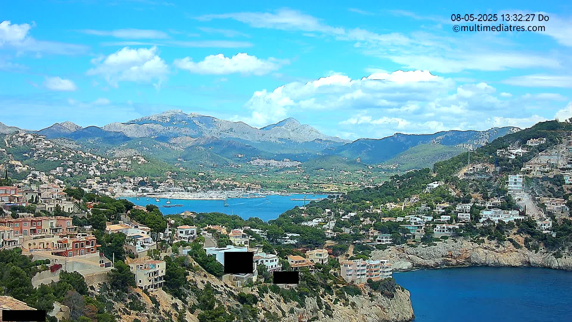 A coastal town nestled on a hillside overlooking a calm bay, with mountains in the background under a partly cloudy sky.