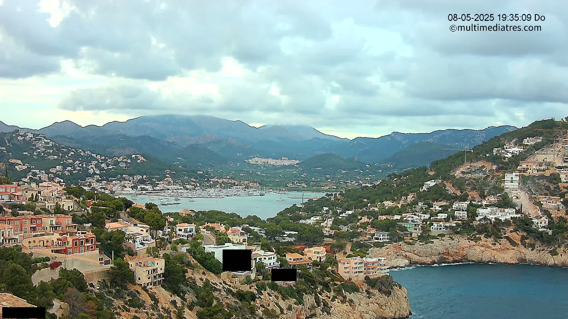 An overcast day reveals a coastal town nestled against a mountainous backdrop, with a harbor filled with boats in the foreground.