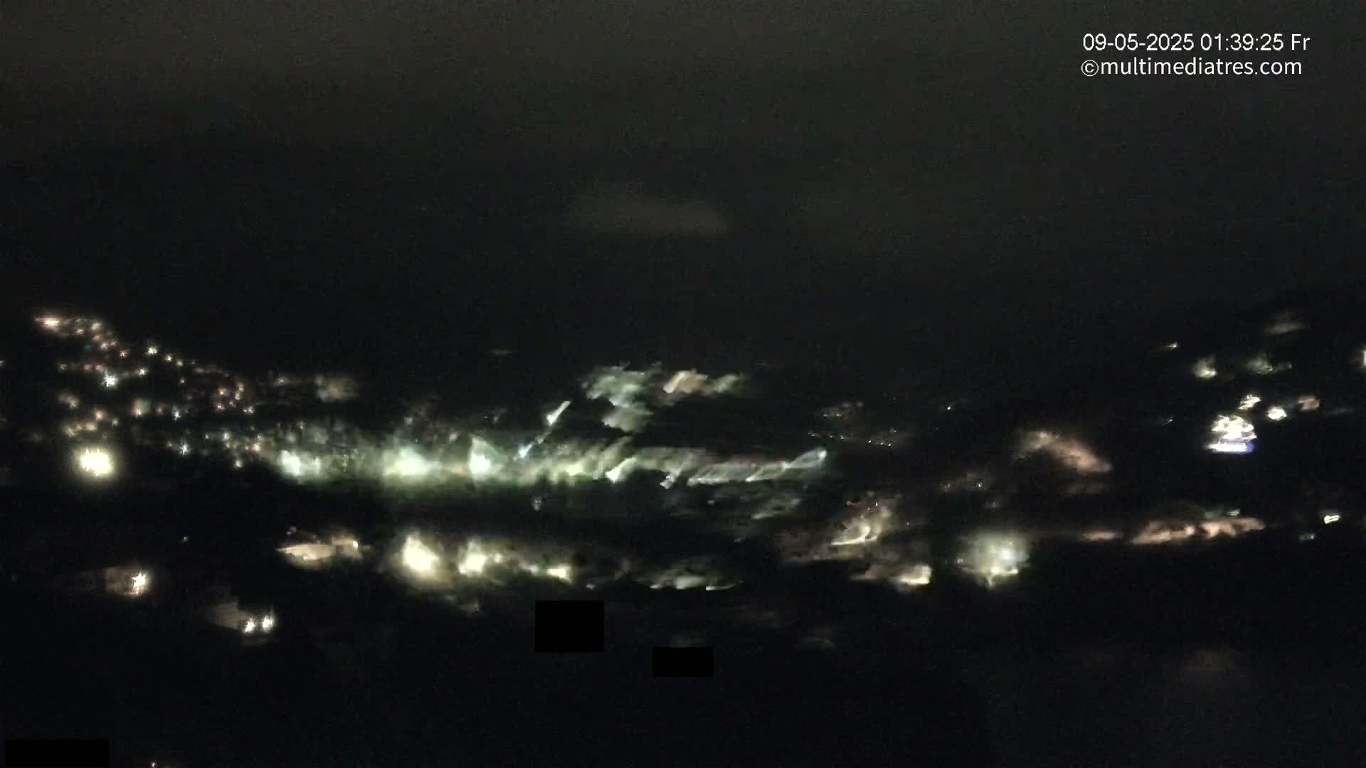 A nighttime aerial view shows a sprawling town illuminated with scattered lights, partially obscured by a hazy or cloudy atmosphere.