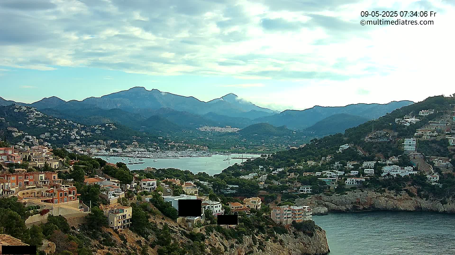 A coastal town nestled in a mountainous region under a partly cloudy sky, features a harbor filled with boats.