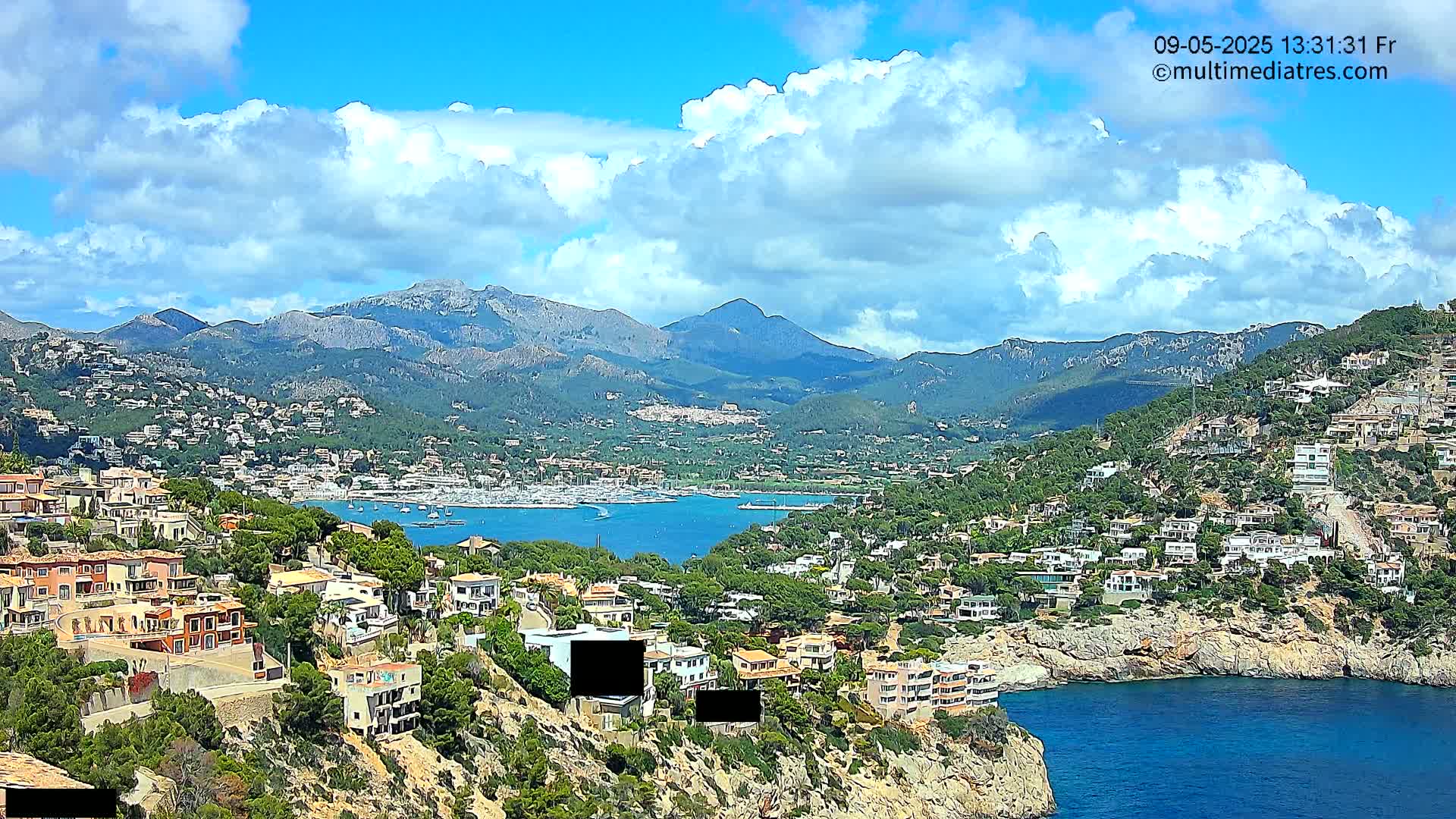 A coastal town nestled in a mountainous region under a partly cloudy sky, overlooks a calm bay filled with boats.