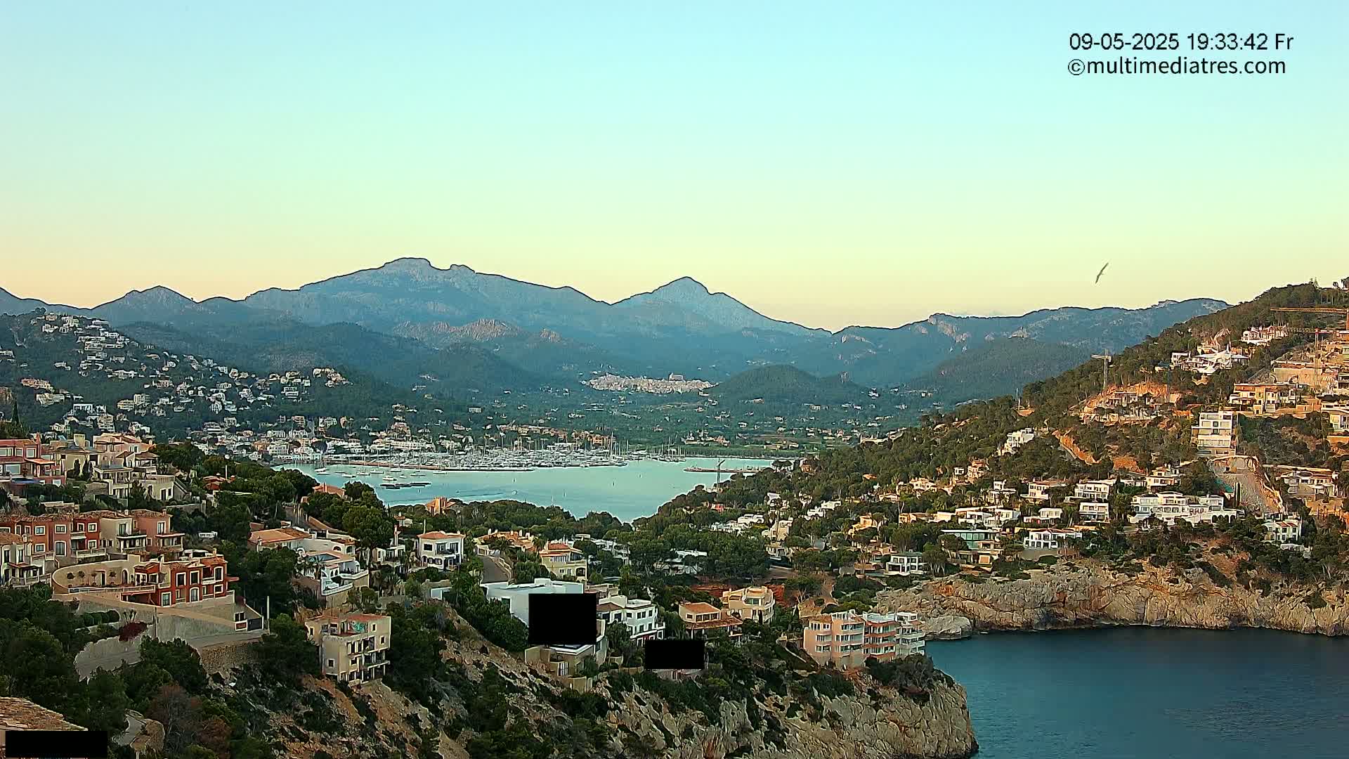 A coastal town nestled in a mountainous region, with a calm bay filled with boats under a clear twilight sky.