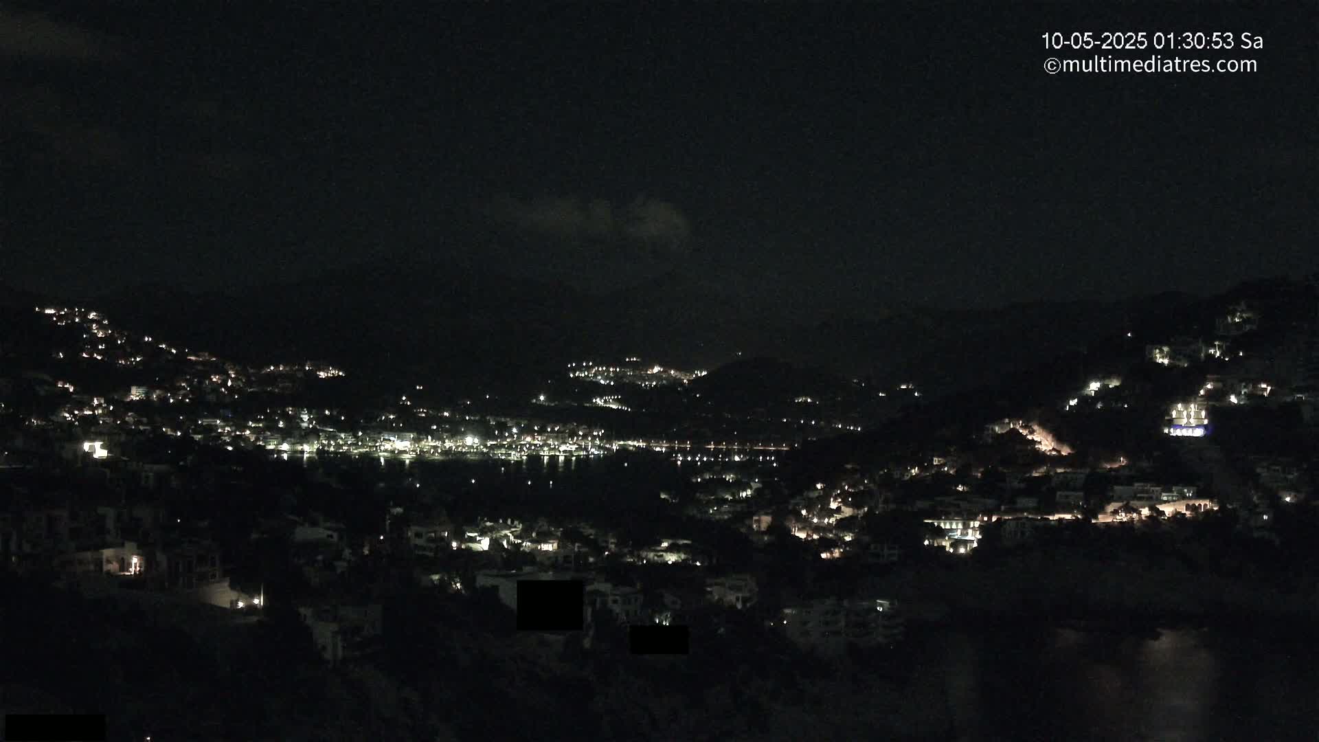 A nighttime view of a coastal town nestled in hills, with scattered lights illuminating the buildings and a dark body of water visible in the distance under a mostly clear sky.