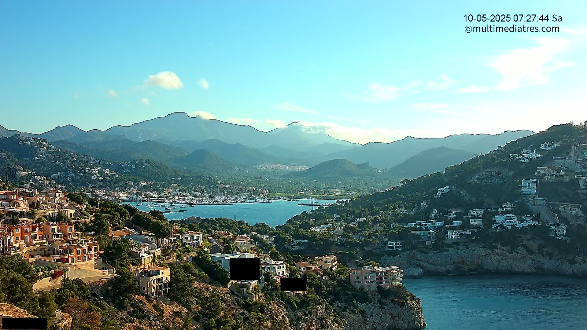 A coastal town nestled on a hillside overlooking a calm bay filled with boats, with mountains in the background under a mostly clear sky.