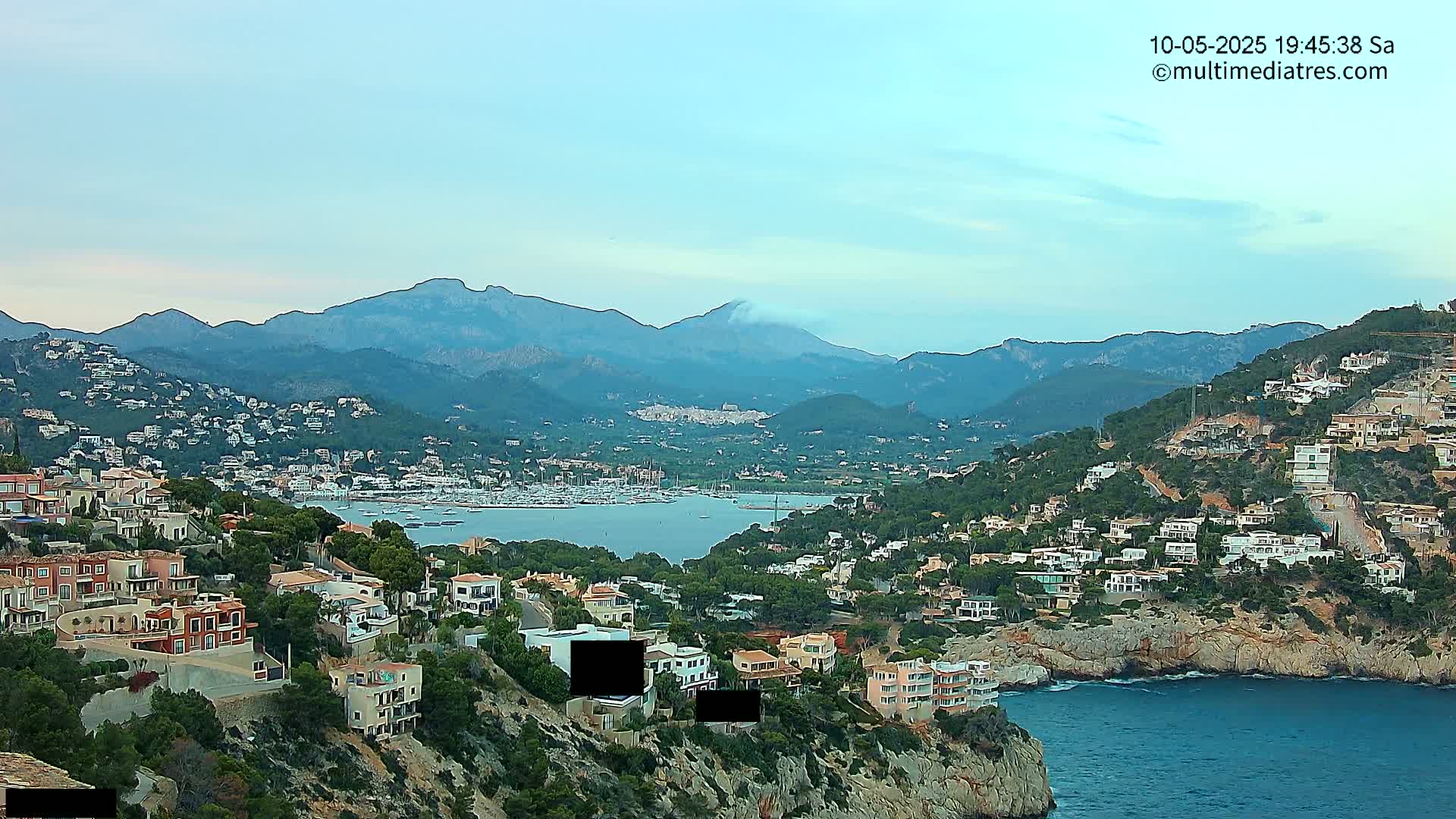 A coastal town nestled into a hillside overlooking a calm bay with a marina, backed by a range of mountains under a mostly clear sky.