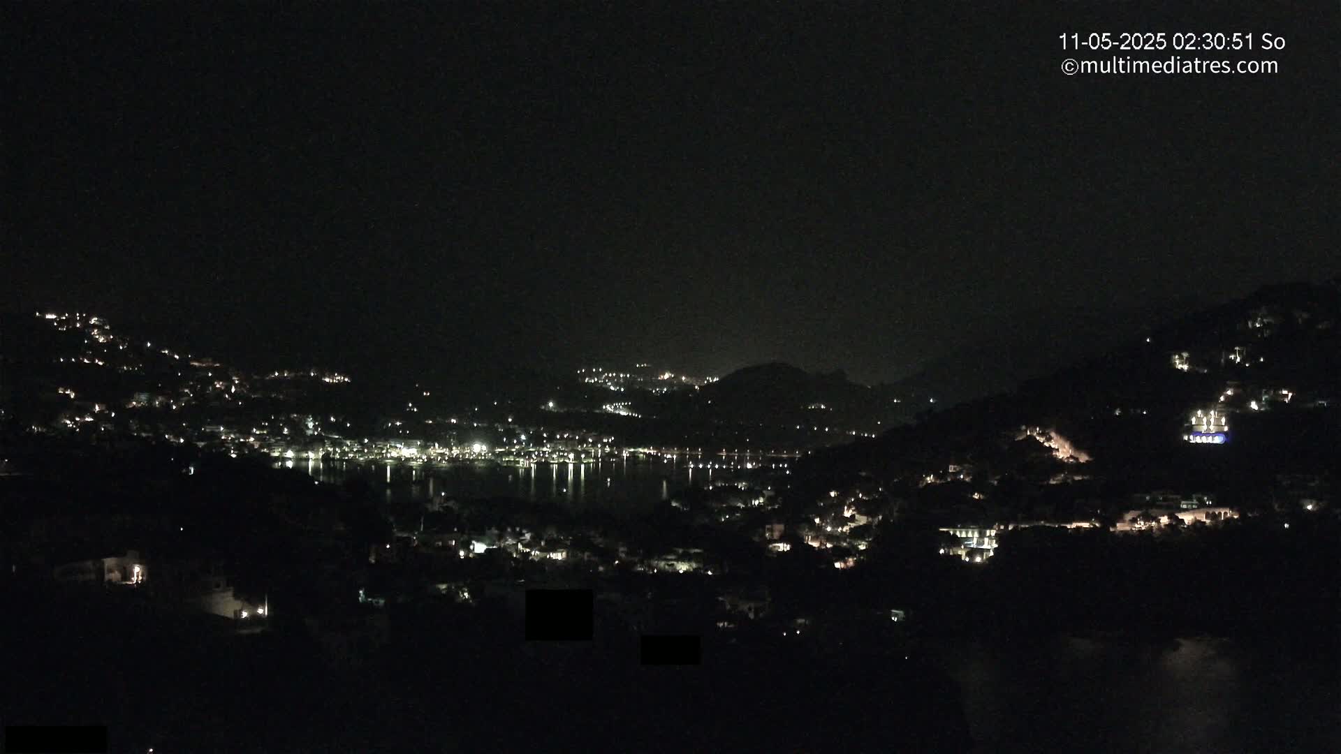 A nighttime view shows a coastal town nestled between hills, its lights twinkling across the dark landscape and reflecting on the water.