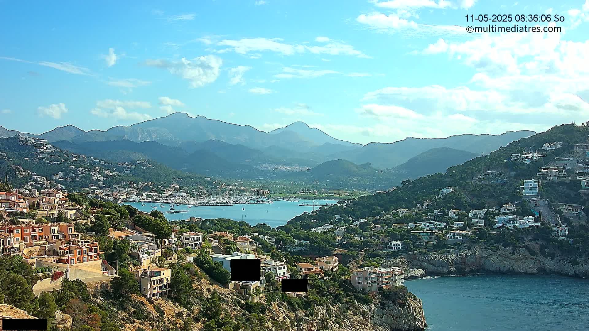 A coastal town nestled on a hillside overlooking a calm bay filled with boats, backed by a mountain range under a mostly clear sky.