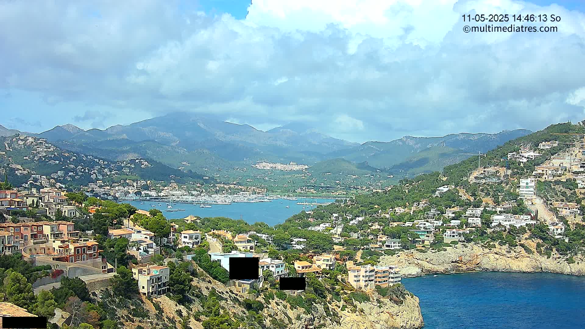 A coastal town nestled amongst hills and mountains overlooking a harbor filled with boats under a partly cloudy sky.