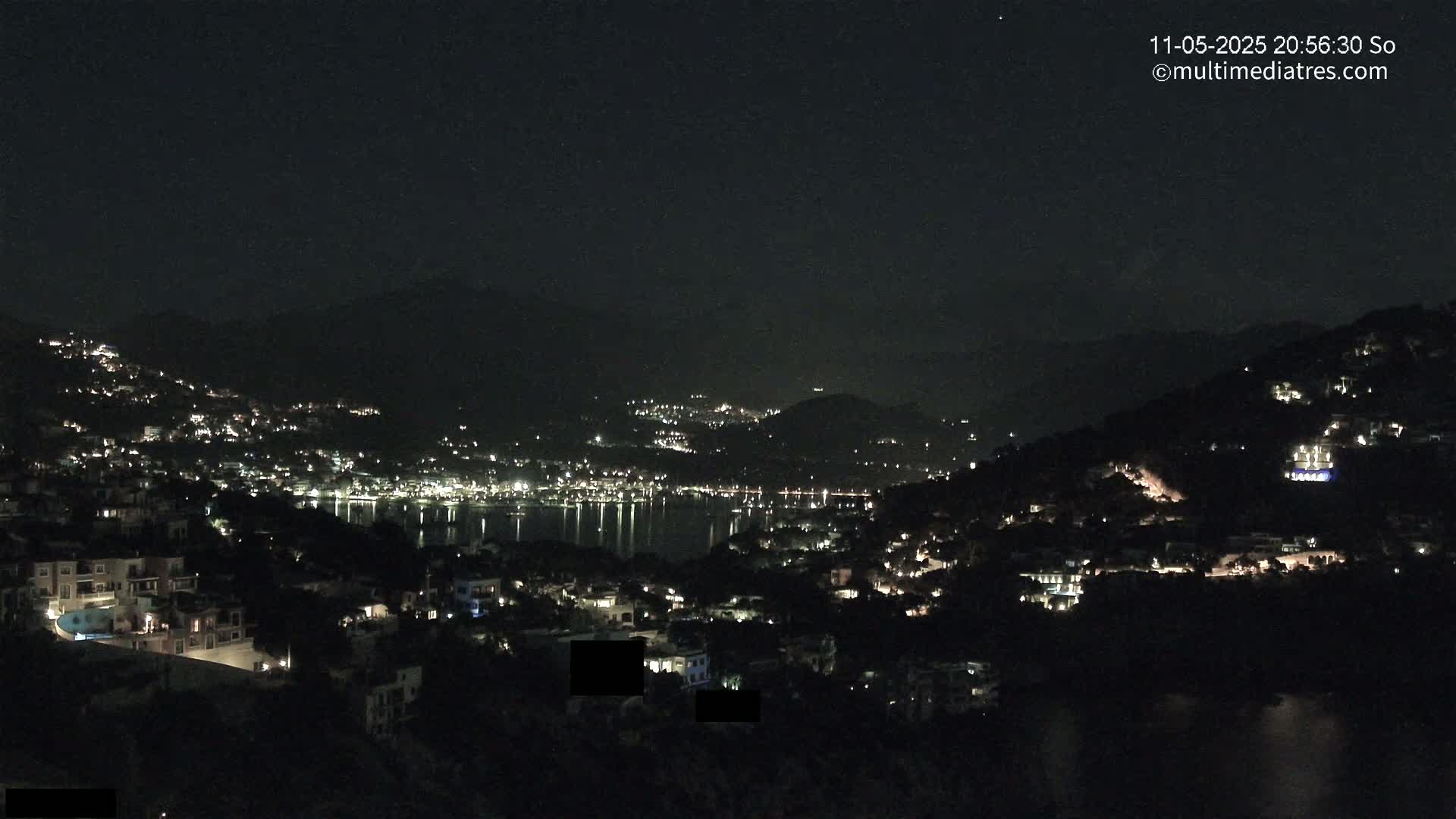 A nighttime view of a coastal town nestled between hills, with its lights reflecting on a calm body of water under a clear, dark sky.