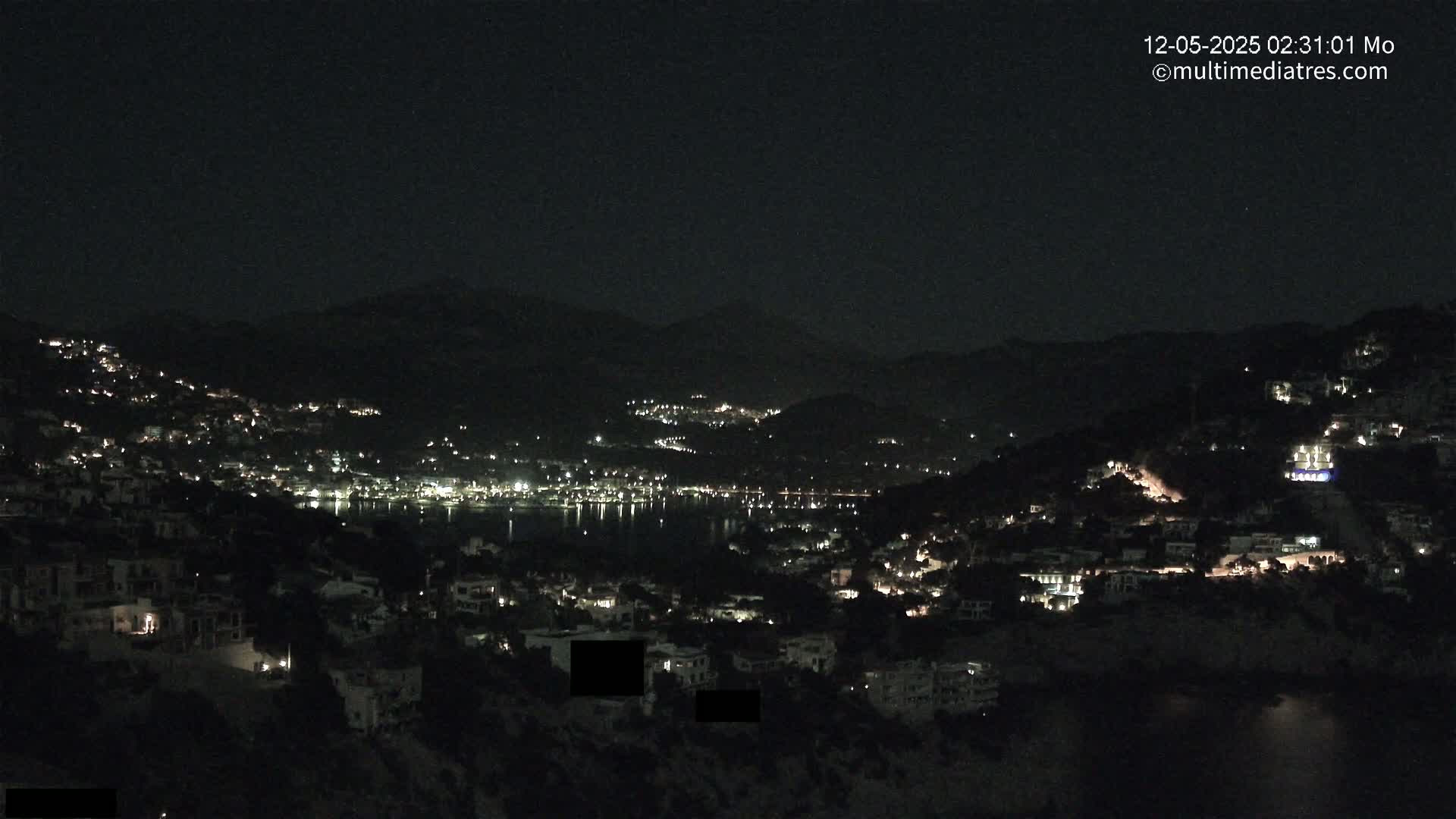 A nighttime view of a coastal town nestled on hillsides, with scattered lights illuminating buildings and a dark body of water reflecting the lights.