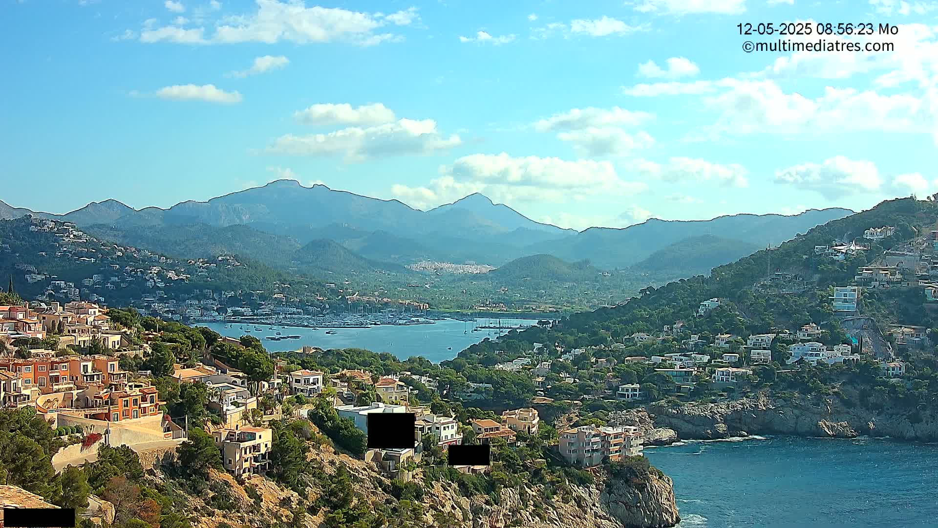 A coastal town nestled on a hillside overlooks a calm bay filled with boats, with mountains in the background under a partly cloudy sky.