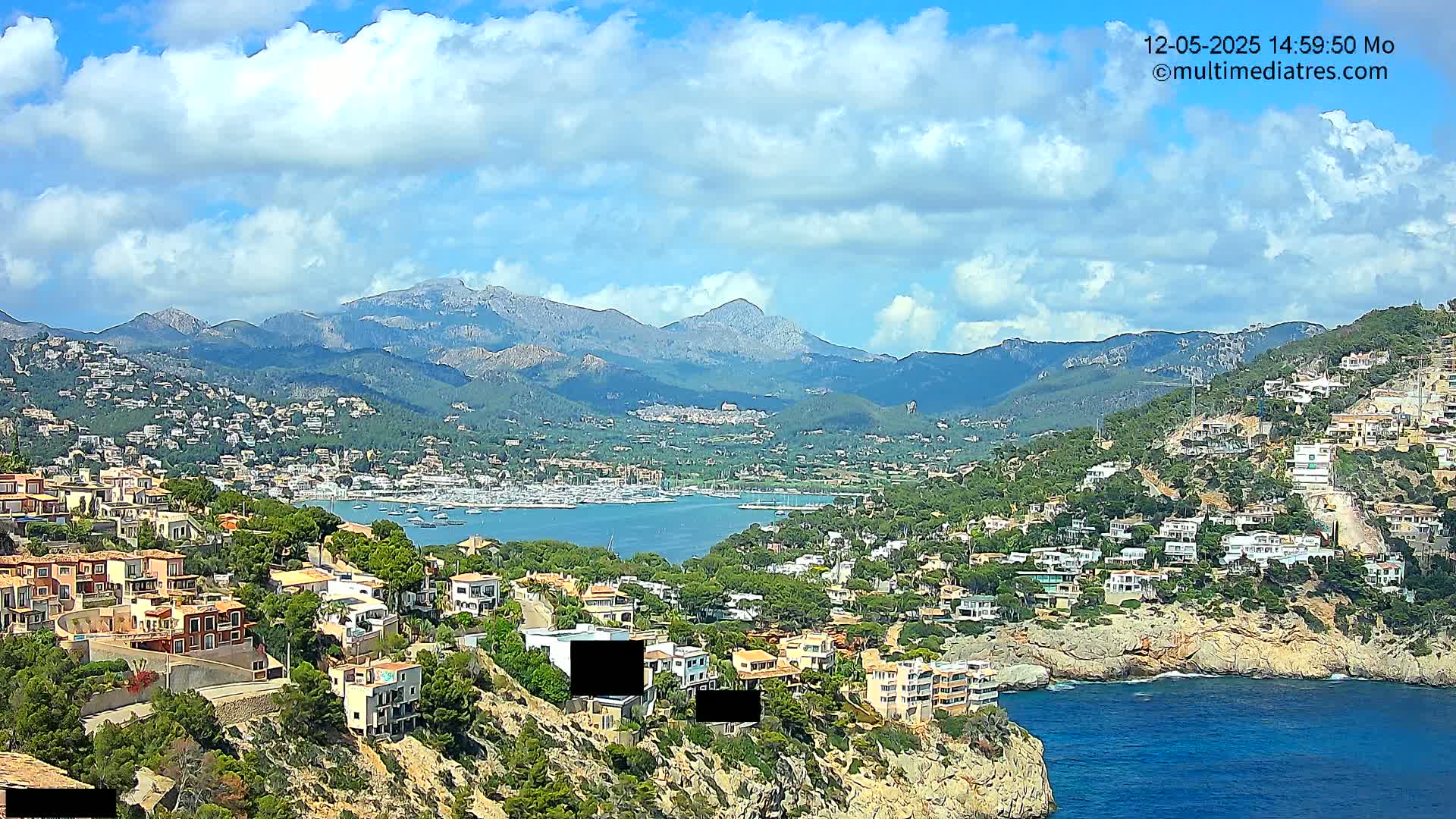 A coastal town nestled on a hillside overlooking a calm bay with numerous boats, backed by mountains under a partly cloudy sky.