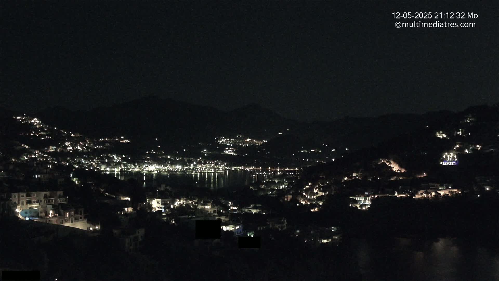 A nighttime view of a coastal town nestled in hills, with numerous lights illuminating buildings and streets, reflecting on the dark water.