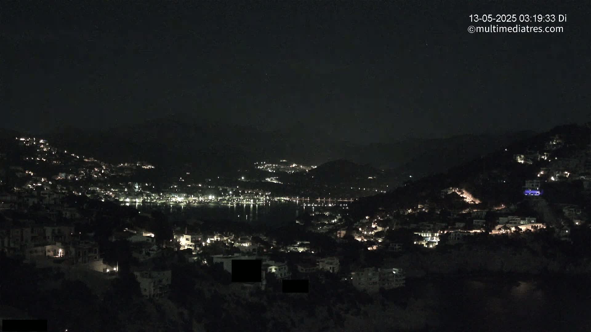 A nighttime view of a coastal town illuminated by many lights, nestled between dark hills under a clear, dark sky.