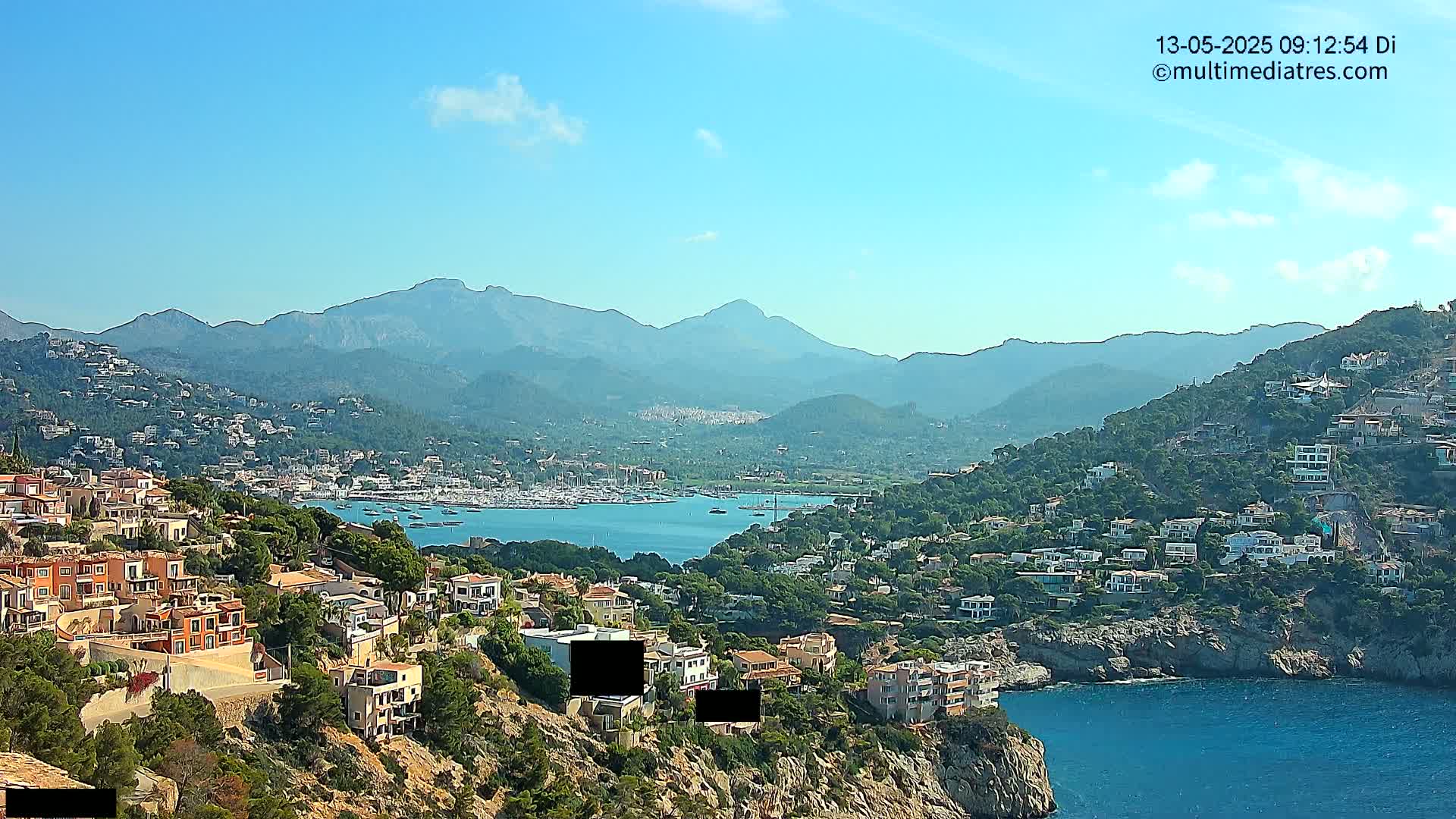 A coastal town nestled against a mountain range under a clear blue sky, with a calm bay filled with boats in the foreground.