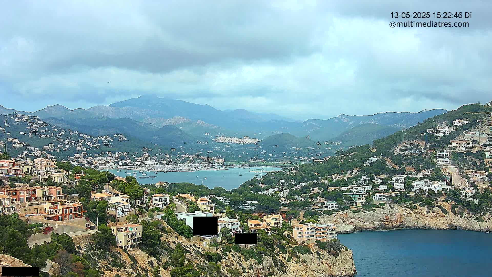 A coastal town nestled in mountainous terrain under a cloudy sky overlooks a calm bay filled with boats.