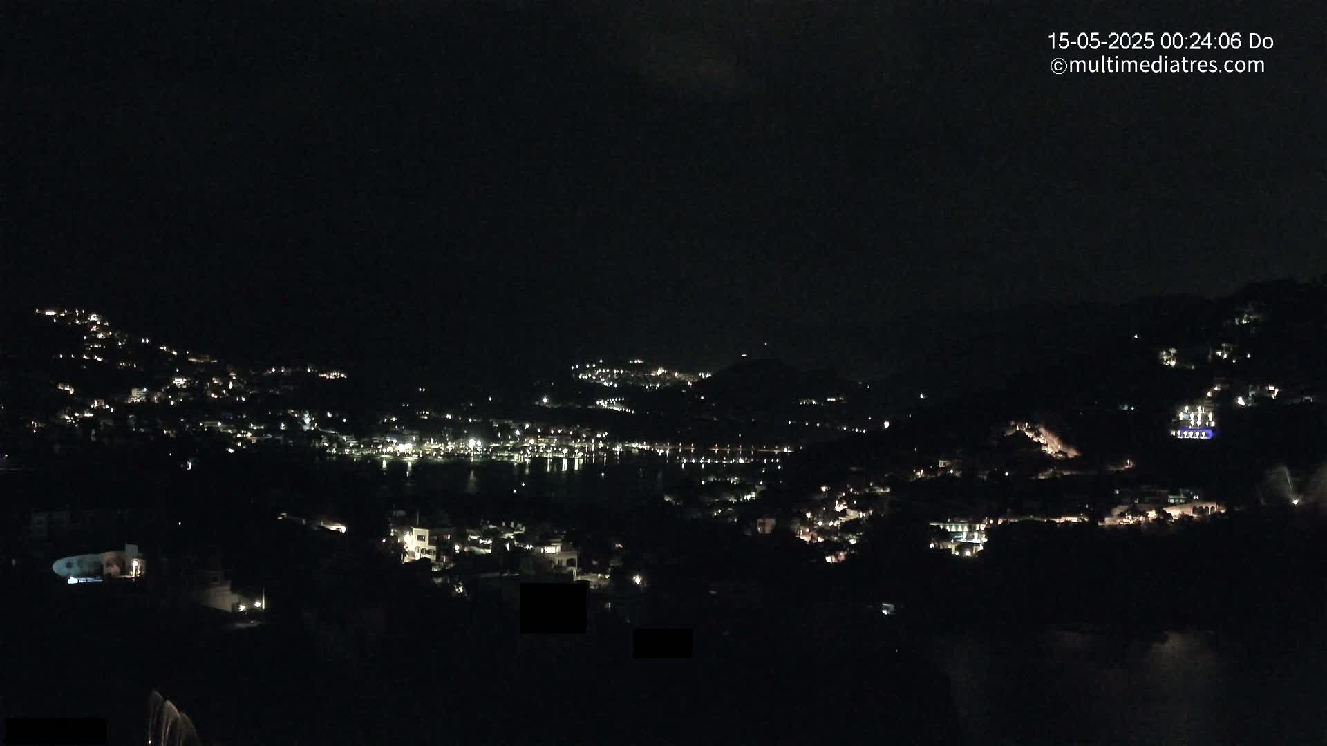 A nighttime view of a coastal town nestled on hillsides, with scattered lights illuminating the buildings and a calm body of water reflecting the lights below.