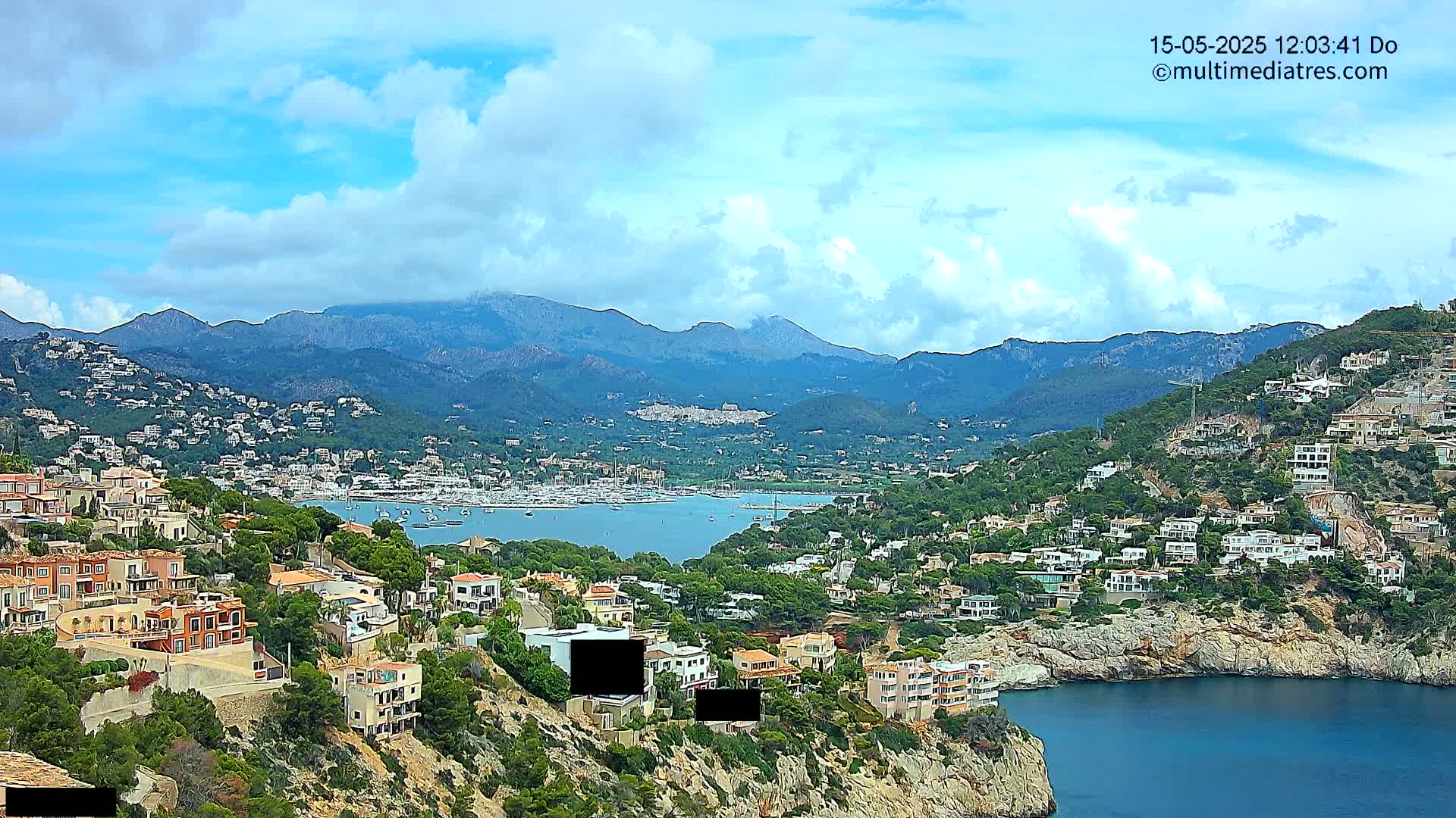 A partly cloudy day overlooks a coastal town nestled among hills and mountains, with a harbor filled with boats visible in the distance.