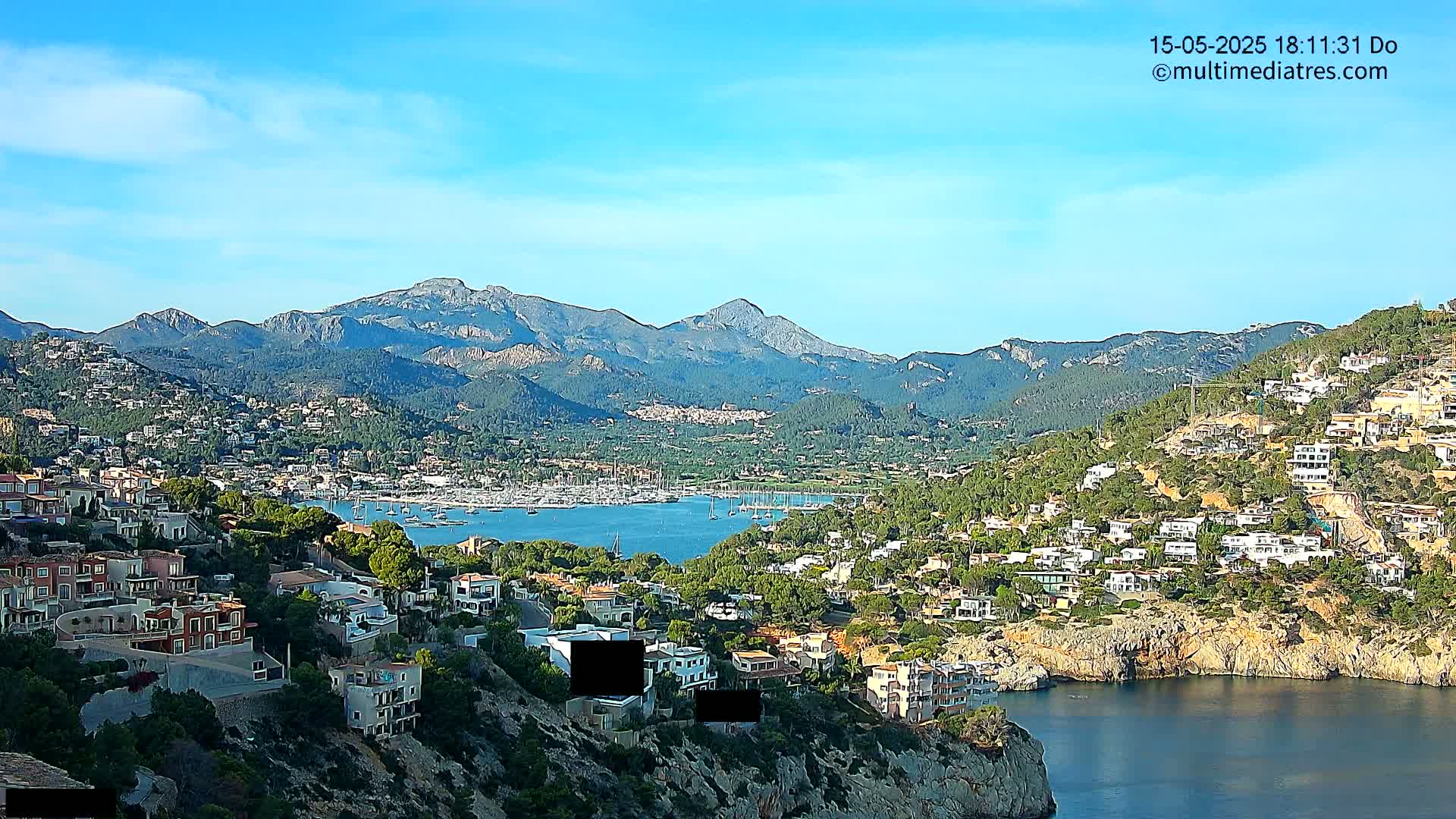 A coastal town nestled among hills and mountains overlooks a calm bay filled with sailboats under a clear blue sky.