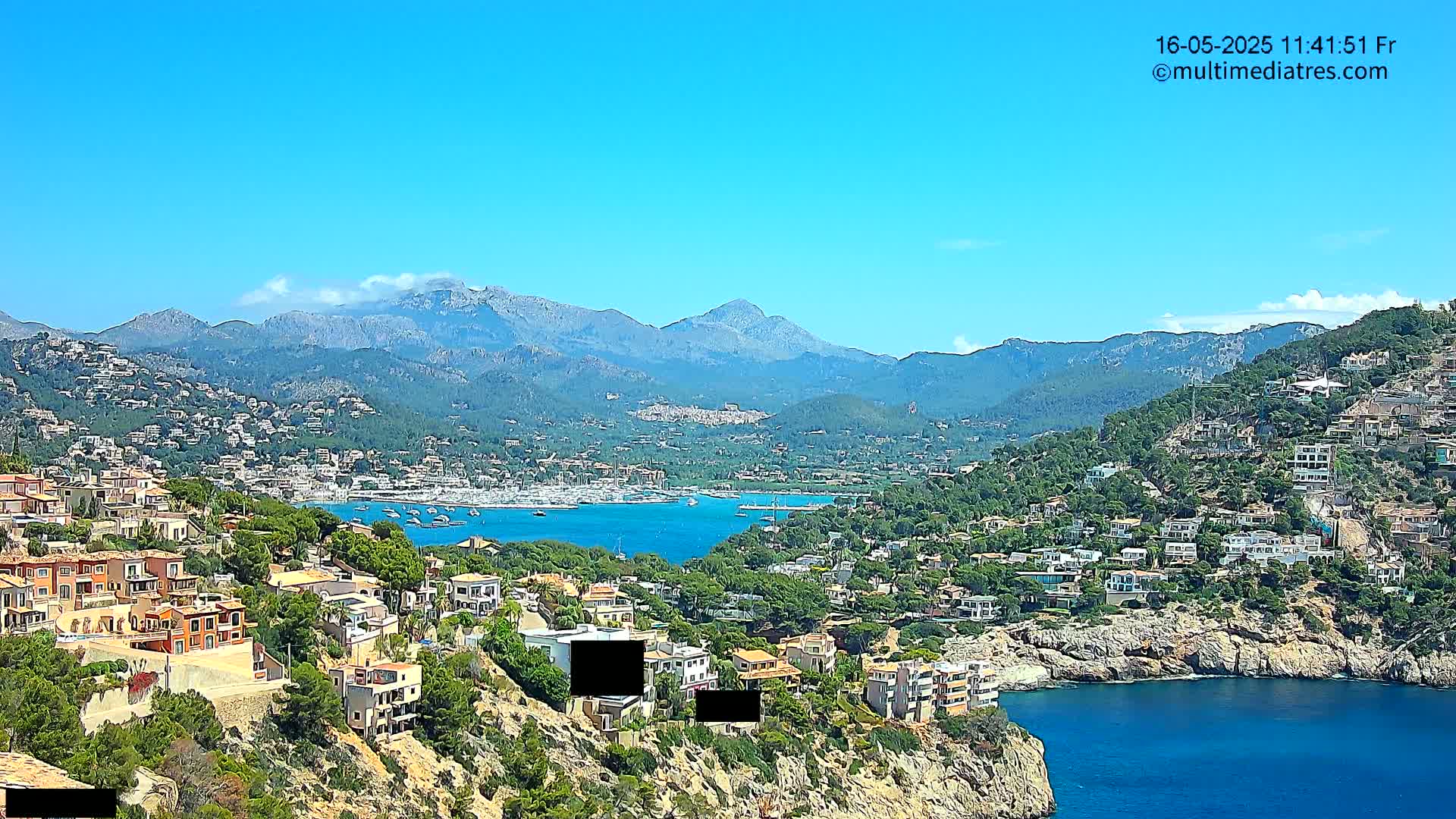 A coastal town nestled on a hillside overlooking a calm bay, with a mountain range in the background under a clear blue sky.