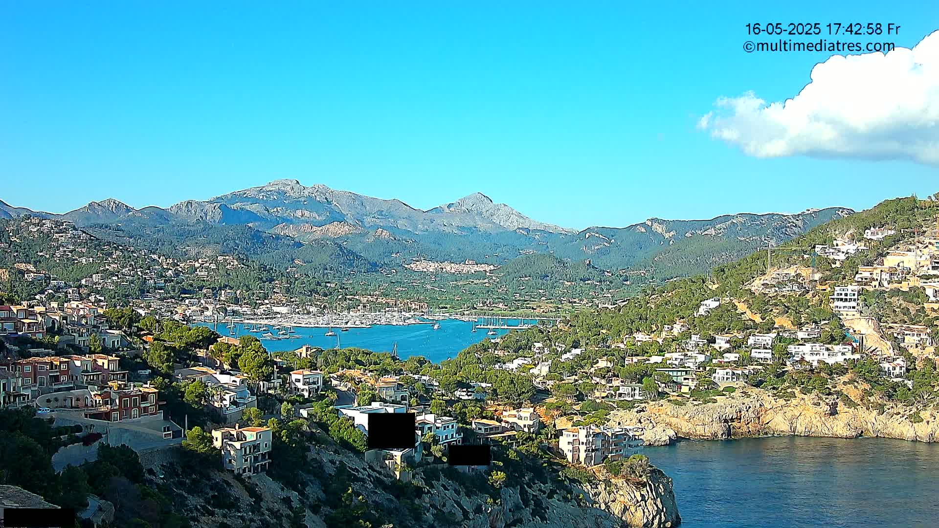 A coastal town nestled against a mountain range under a clear blue sky with a few scattered clouds, features a harbor filled with boats.