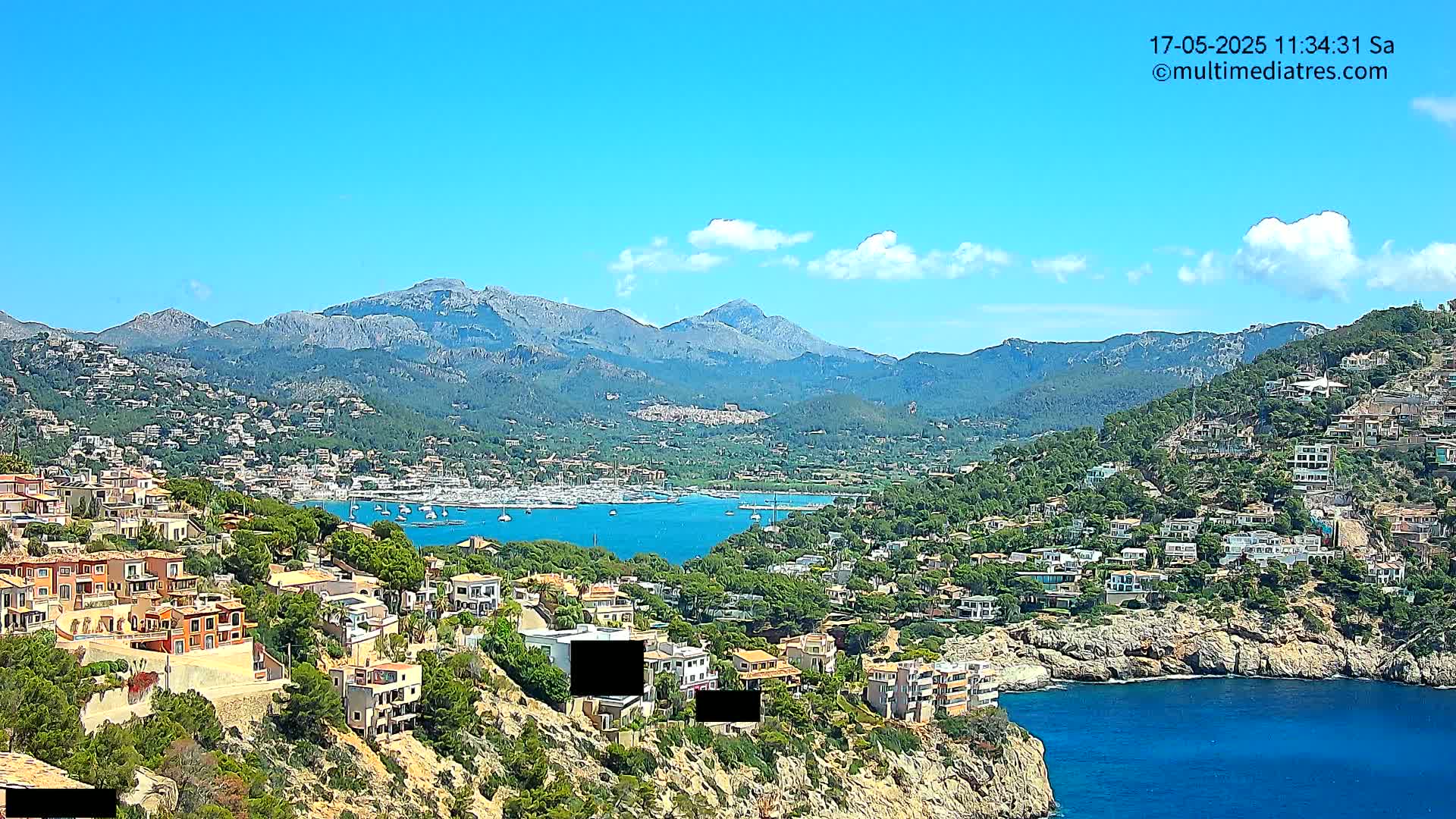 A sunny day overlooking a coastal town nestled in mountains beside a calm blue bay with numerous boats.