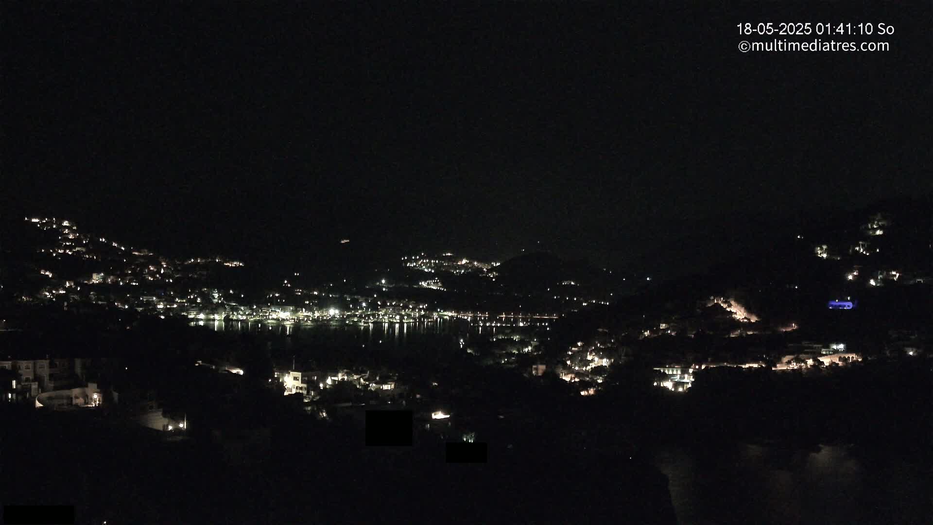 A nighttime view of a town nestled on hillsides surrounding a dark body of water, illuminated by scattered lights.