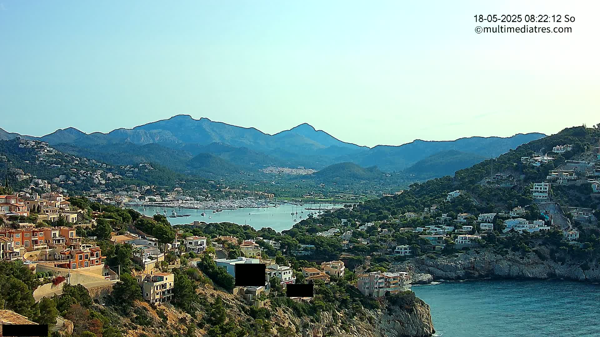 A coastal town nestled against a mountain range under a clear sky, with a marina filled with boats and numerous houses dotting the hillside.