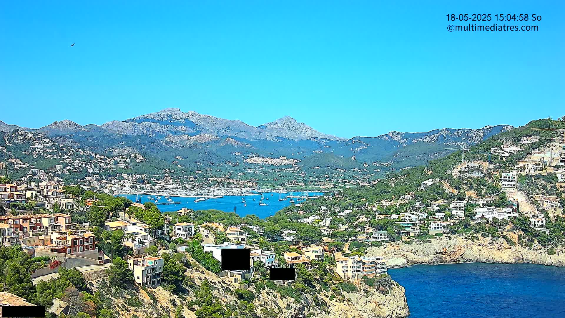 A coastal town nestled on a hillside overlooking a bay filled with sailboats and backed by a mountain range under a clear blue sky.
