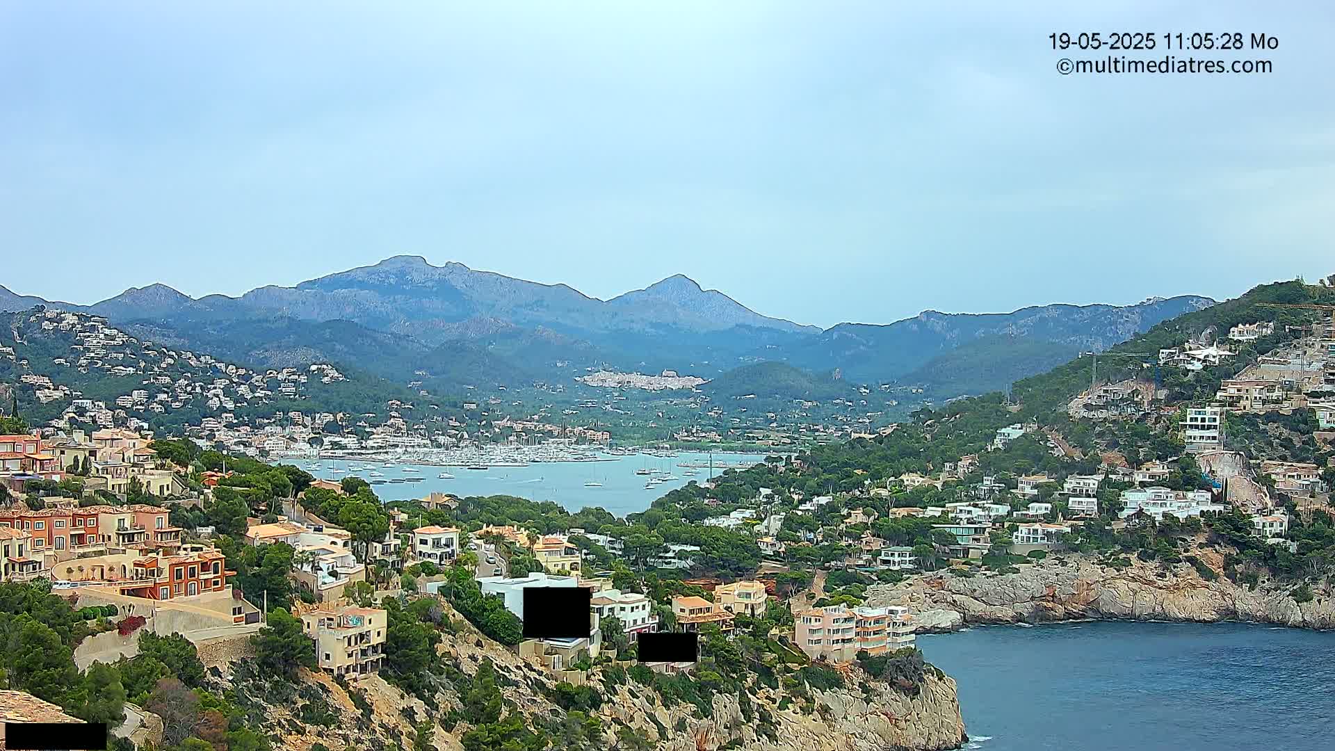 A coastal town nestled between rocky hills and a calm bay, under a mostly clear sky, features many houses, boats in the water and mountains in the background.